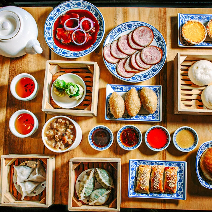 Chinese dim sum spread with dumplings, siomai, steamed buns, spring rolls, sauces, and tea served on a wooden table.