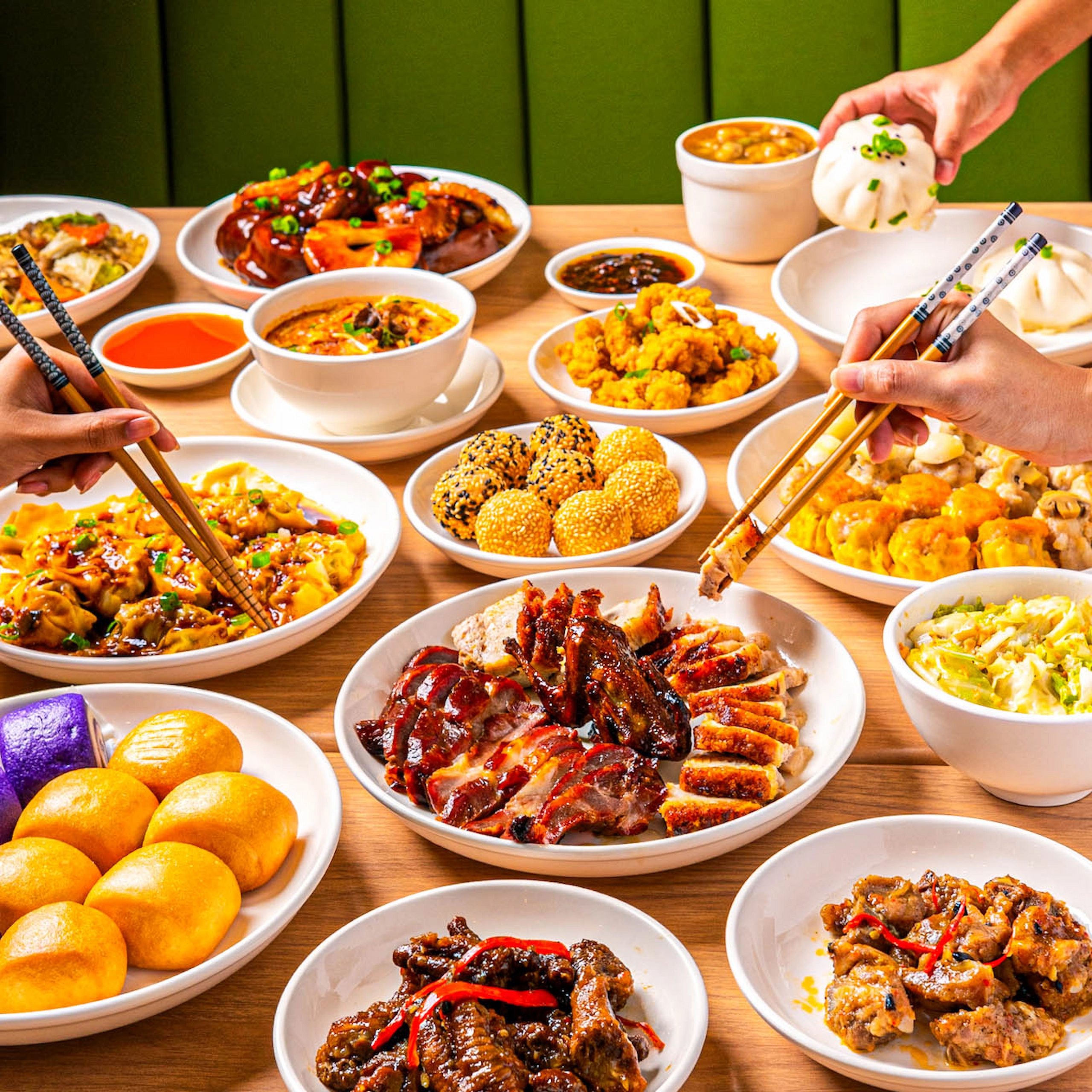 Table filled with Chinese dishes including dim sum, roast meats, dumplings, fried dishes, and noodles, as diners use chopsticks to share the meal.