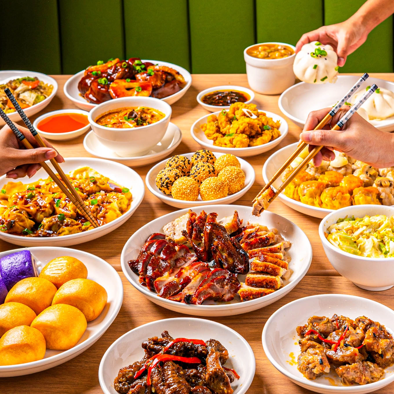 Table filled with Chinese dishes including dim sum, roast meats, dumplings, fried dishes, and noodles, as diners use chopsticks to share the meal.