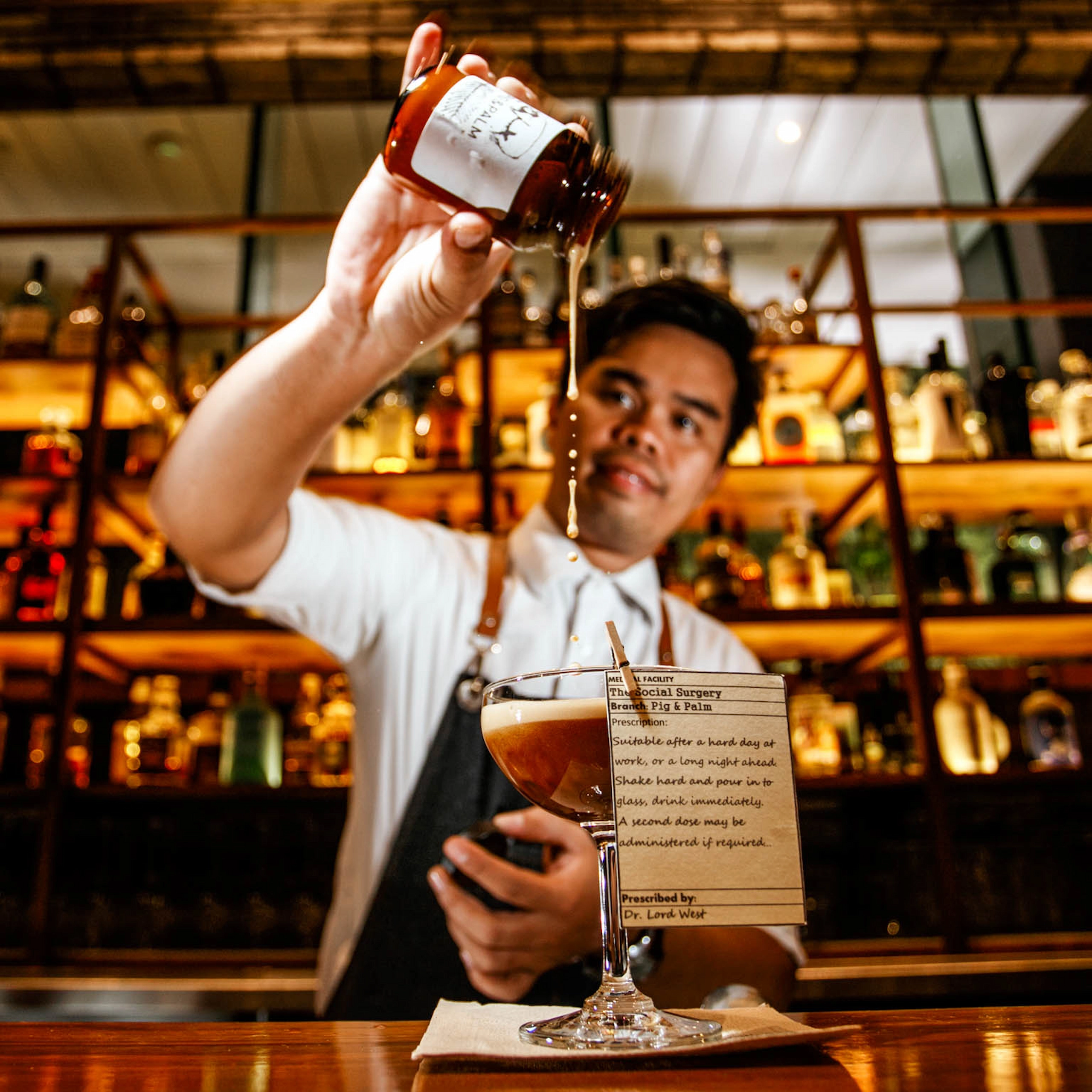 Bartender at The Pig & Palm in Cebu City pouring syrup into a cocktail, with a bar shelf of liquor bottles in the background.