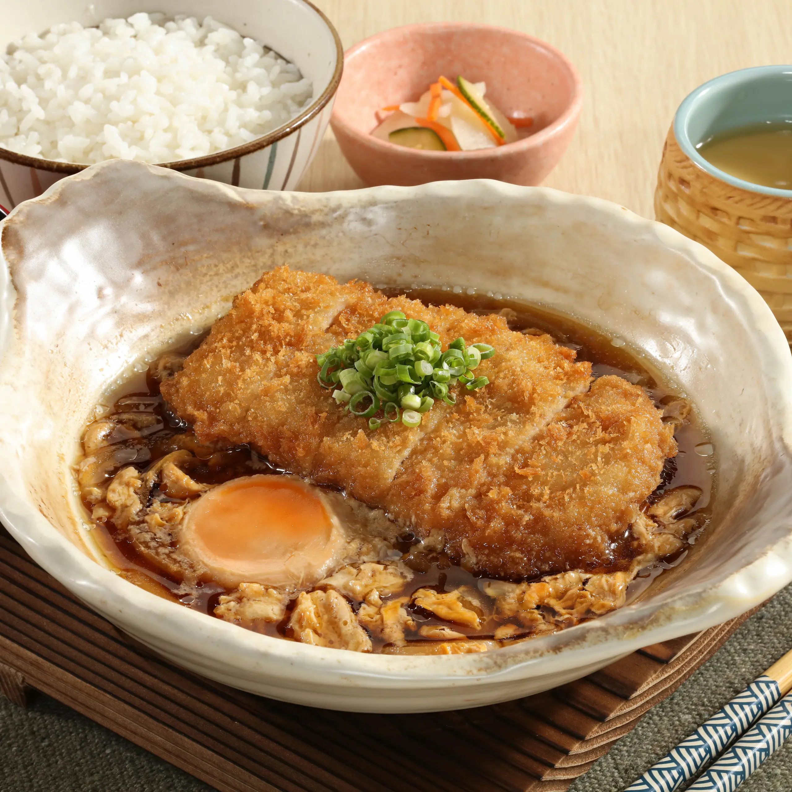 Pork Loin Katsu Tamagotoji served in savory broth with egg and chopped green onions, accompanied by steamed white rice, pickled vegetables, and a cup of tea.