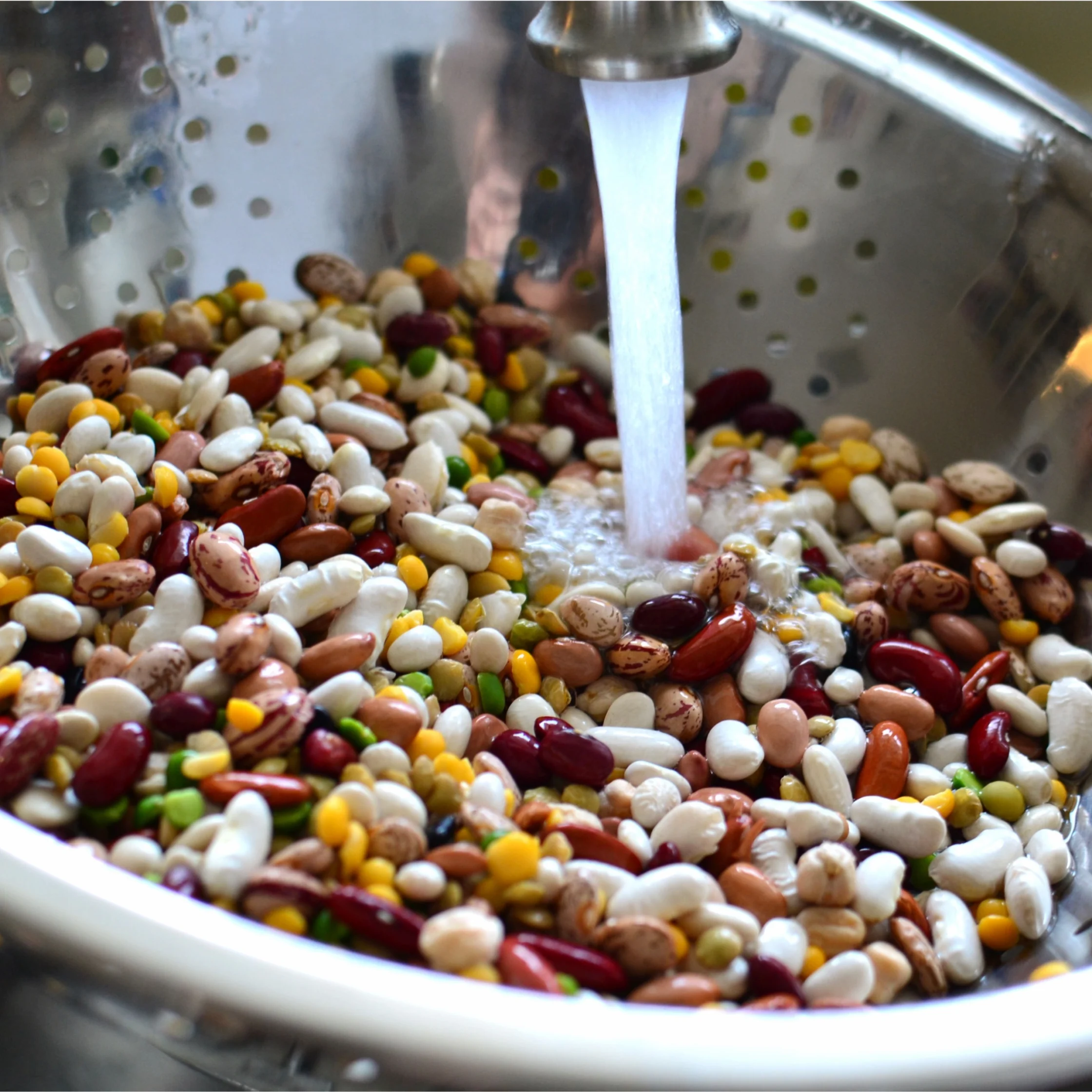 Colander filled with mixed dried beans being rinsed under running water from a faucet