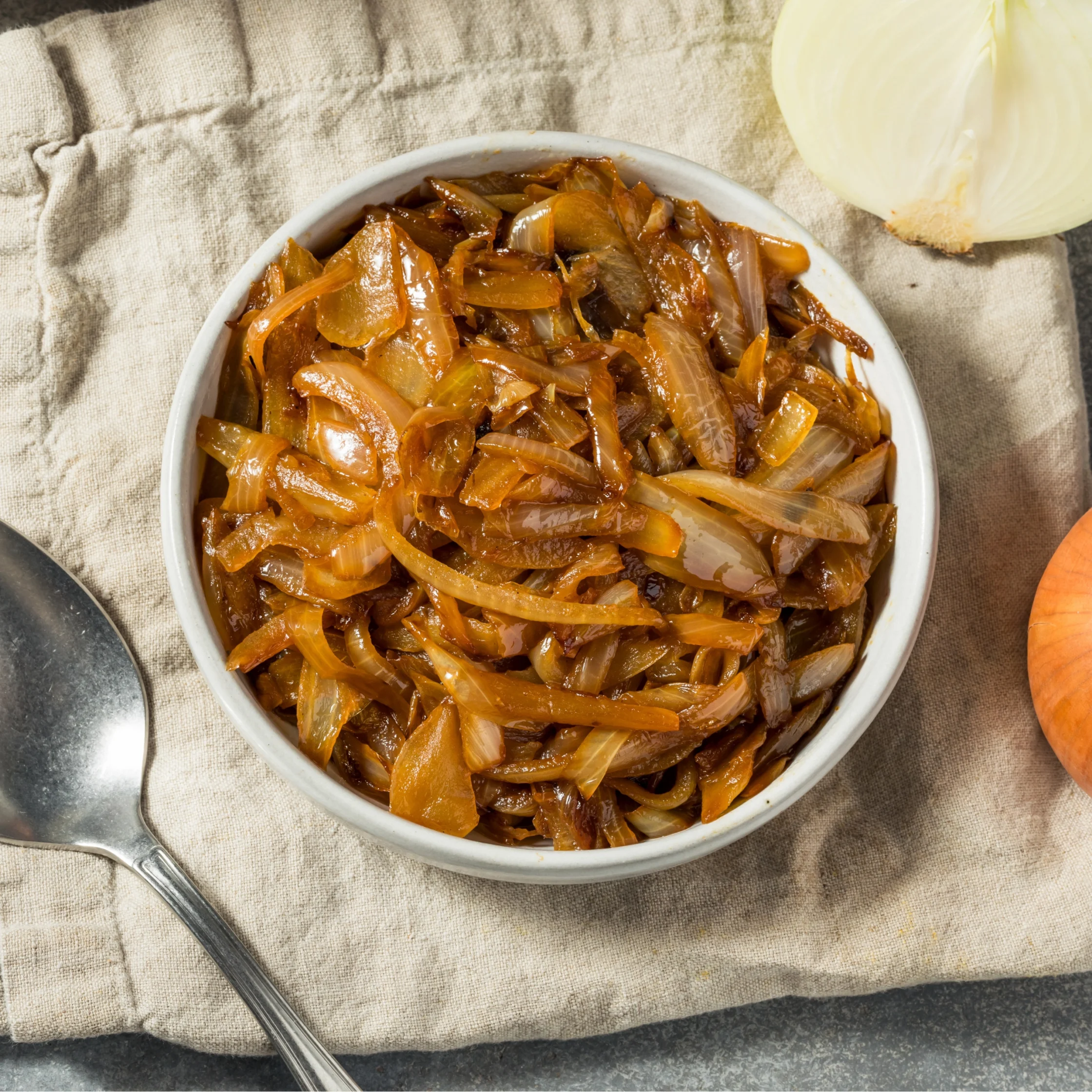 White bowl filled with caramelized onions on a beige linen cloth with a spoon nearby.