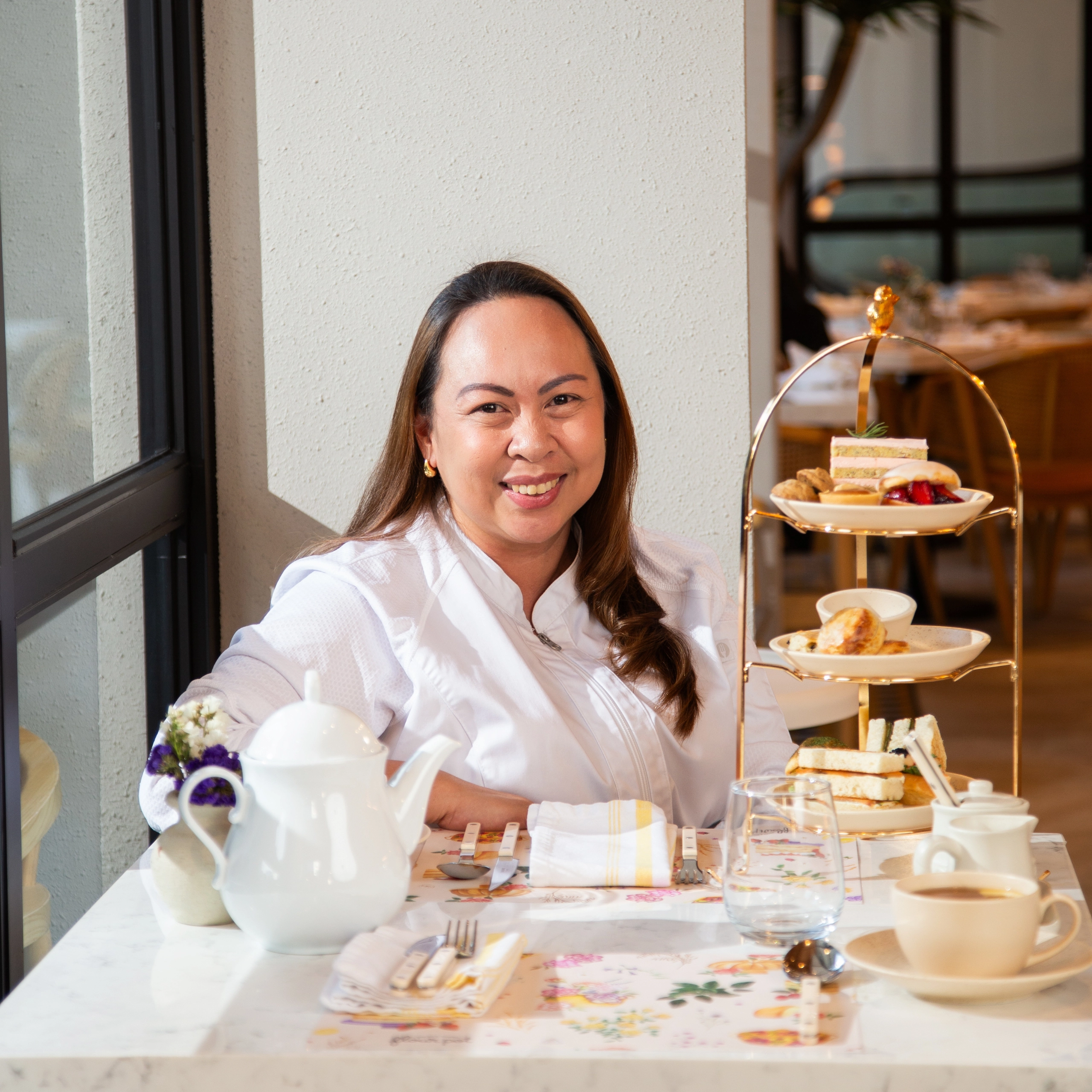 Smiling woman in a white blouse seated at a cafe table with a three-tier stand of pastries and sandwiches nearby. Chef Rhea SyCip of Flour Pot