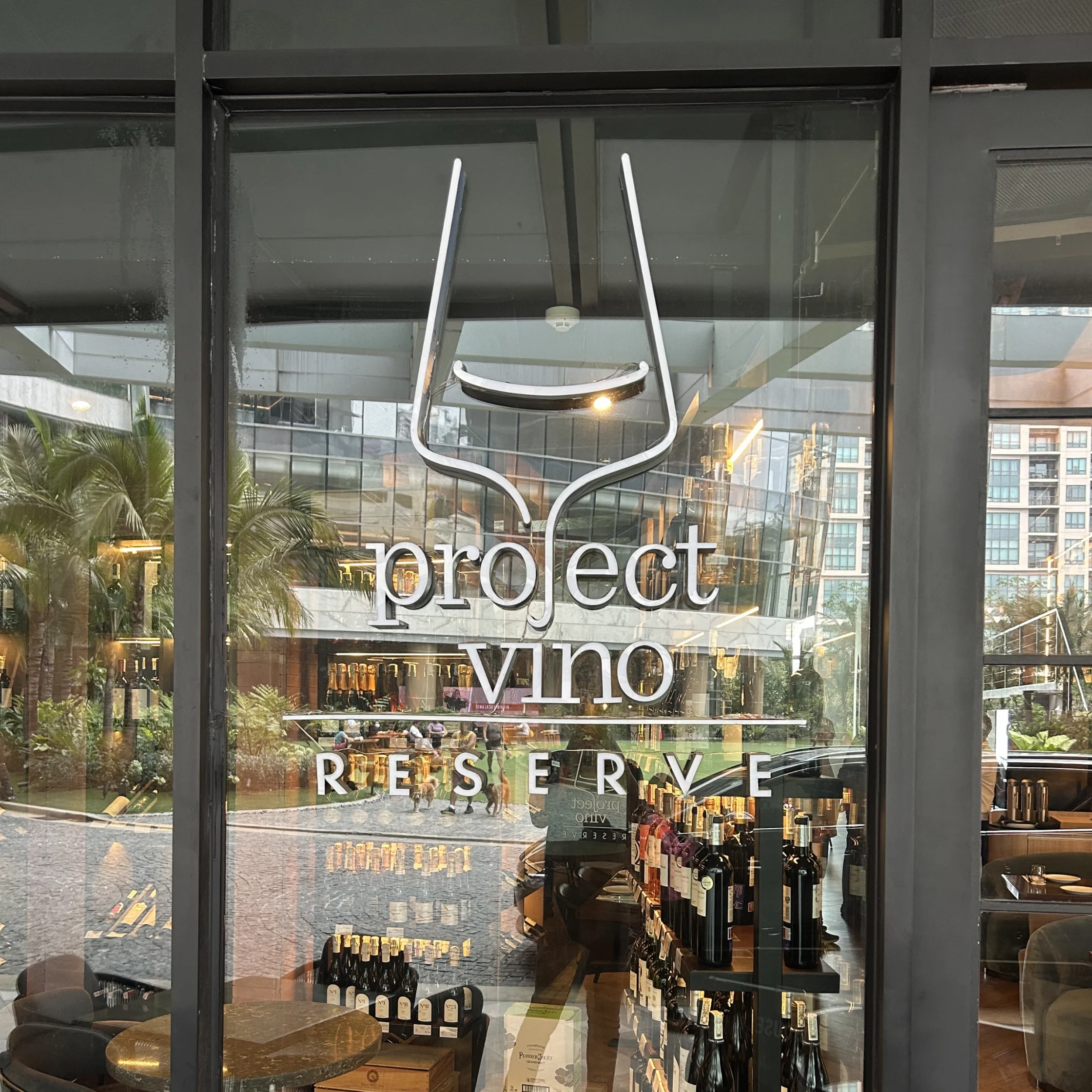 Glass storefront window with a white logo reading 'Project Vino Reserve' and a stylized wine glass, with wine bottles visible inside and reflections of the outside area.