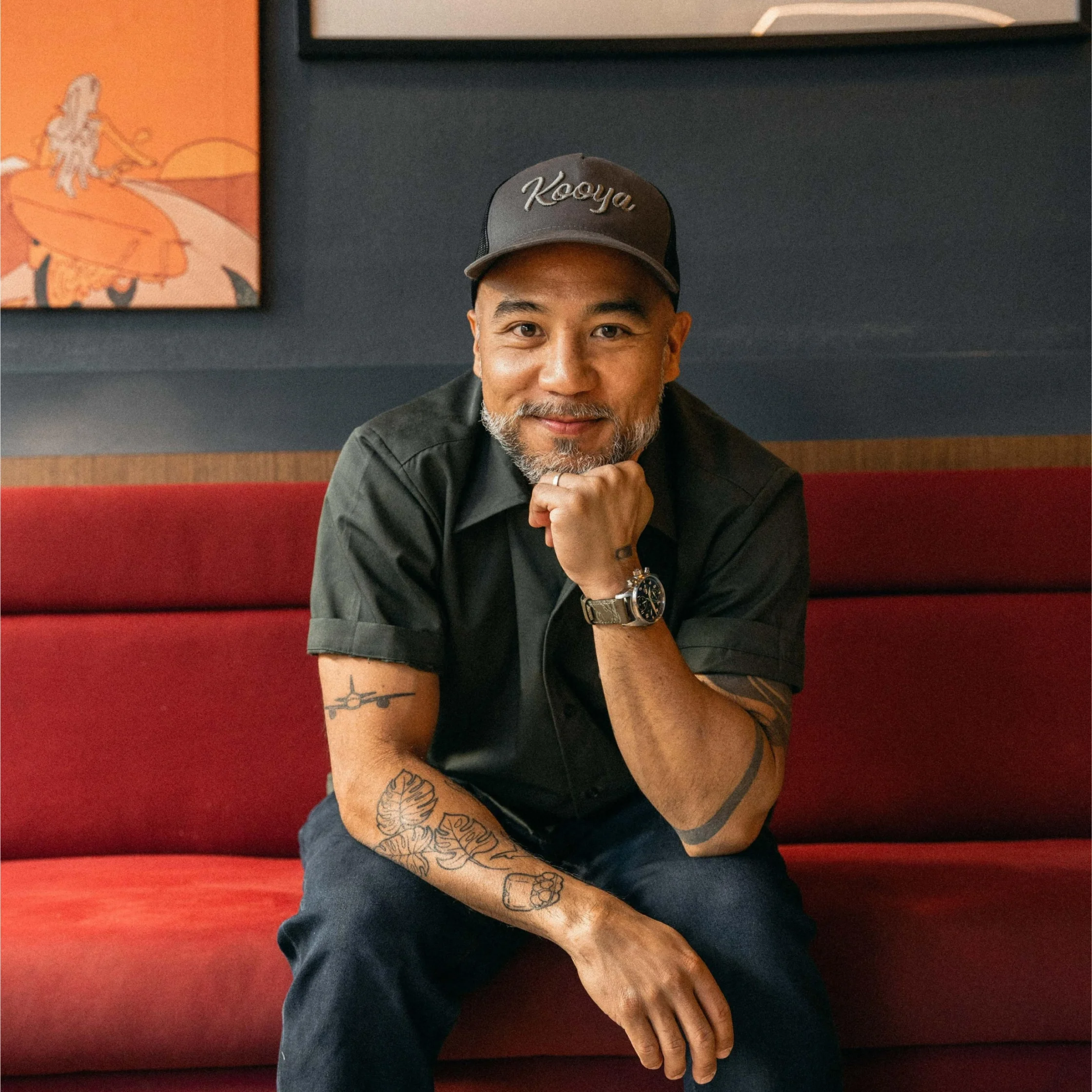 Smiling man with a gray beard wearing a cap and black shirt sits on a red booth, showing tattoos on his arms and a wristwatch.