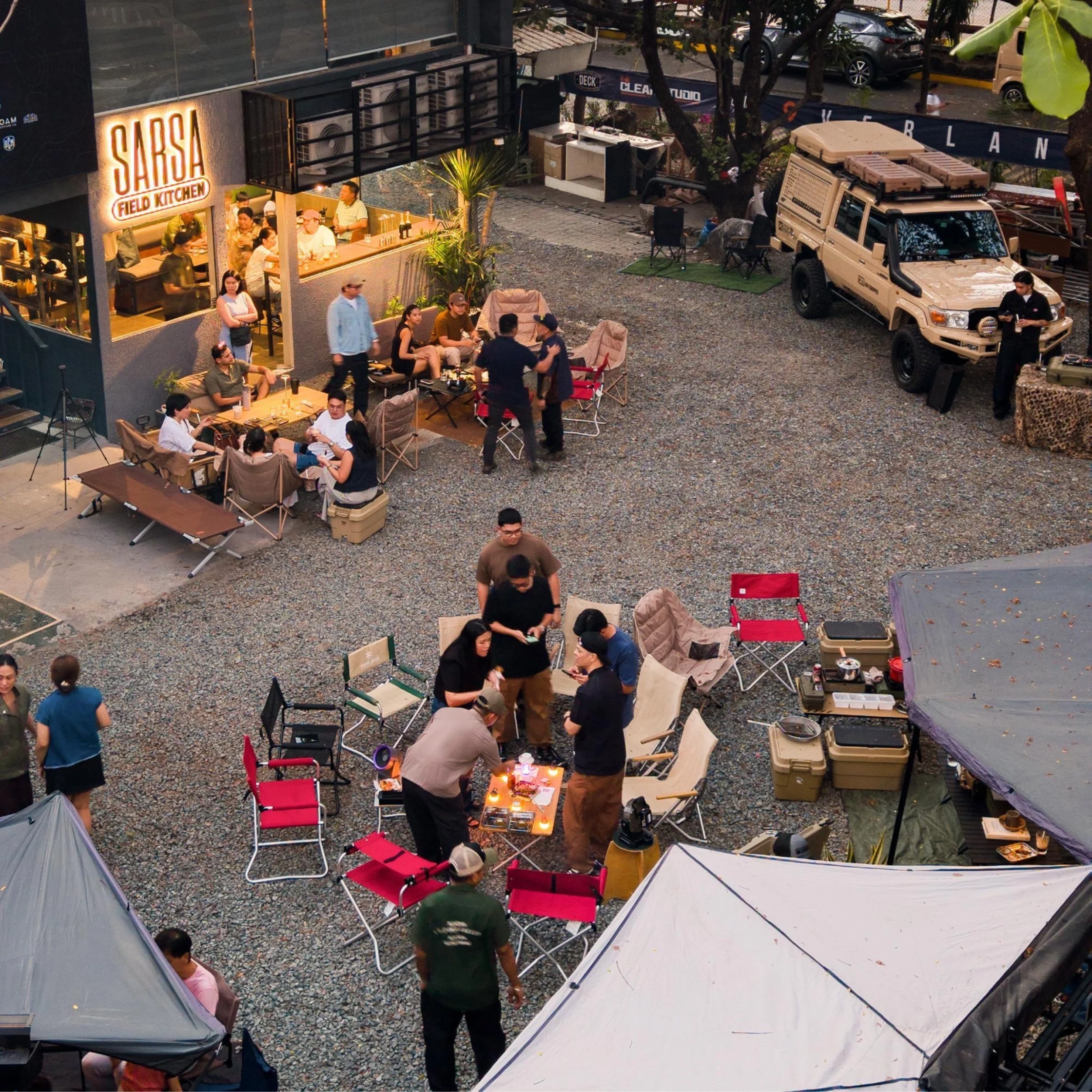 Outdoor group of people socializing at a casual yard party with tents and folding chairs nearby a restaurant exterior at dusk.