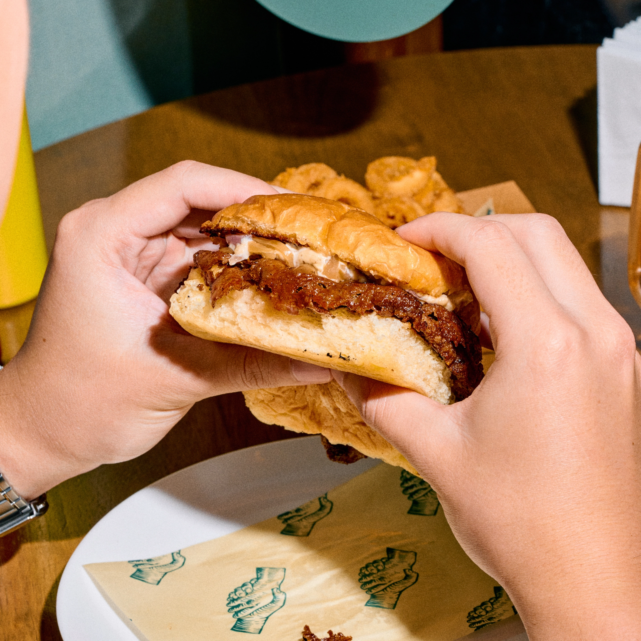 Close-up of hands holding a partially eaten beef burger with melted cheese and sauce, over a plate with a paper wrapper.