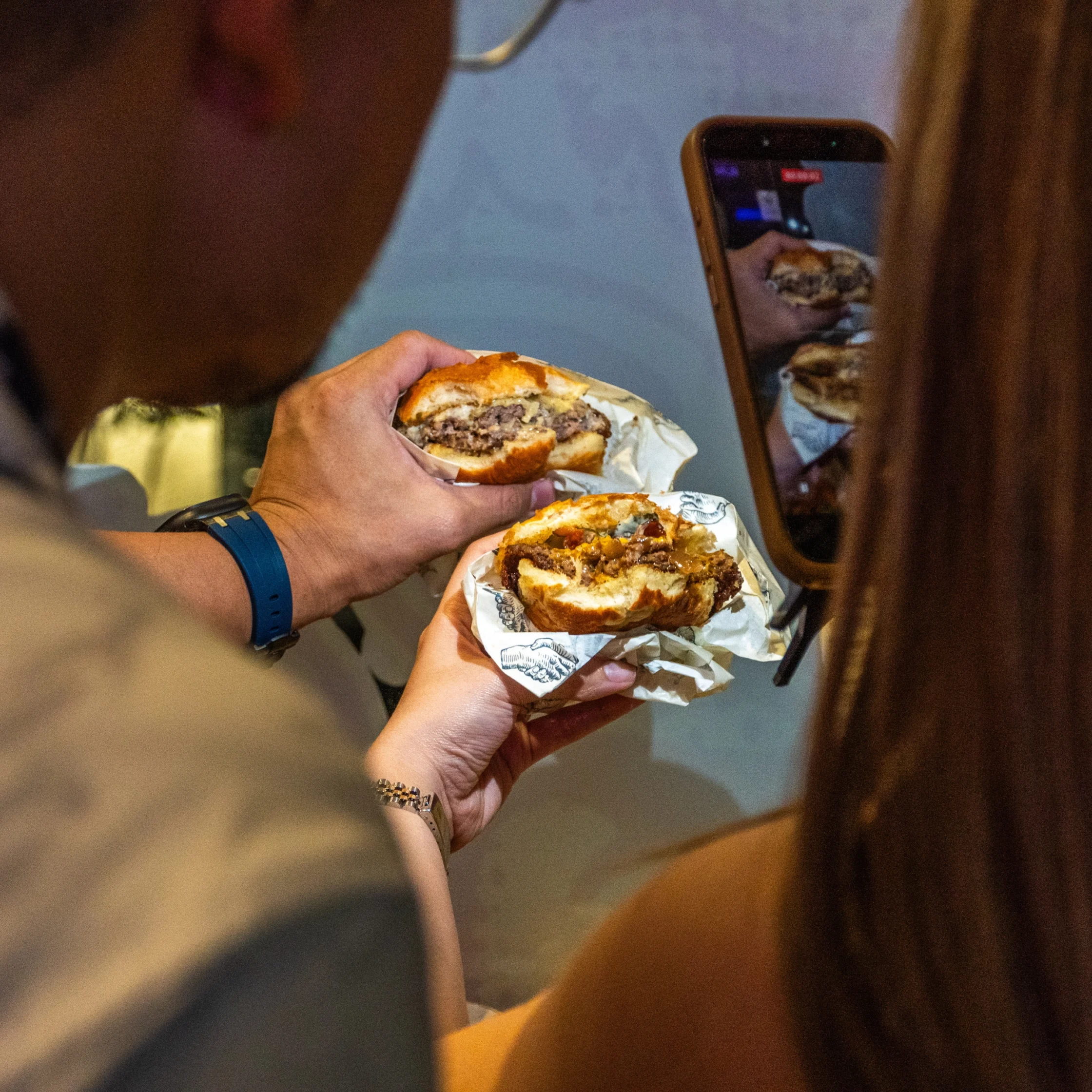 Person holding a burger wrapped in paper while filming or photographing it with a smartphone, reflection of the burger visible on the screen