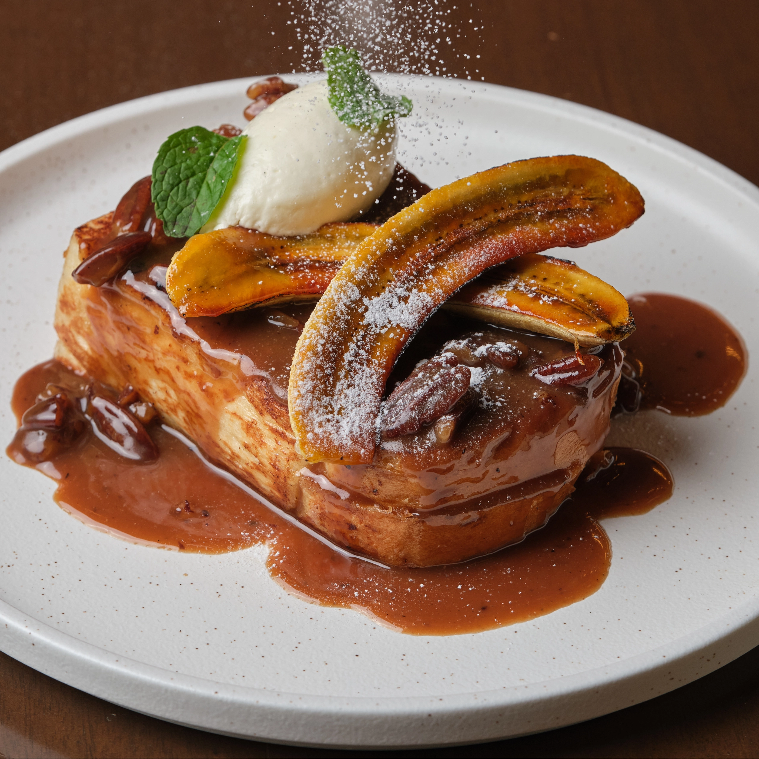 Dessert plate featuring caramel-coated pastry with banana slices, a scoop of vanilla ice cream, mint leaf, and a dusting of powdered sugar