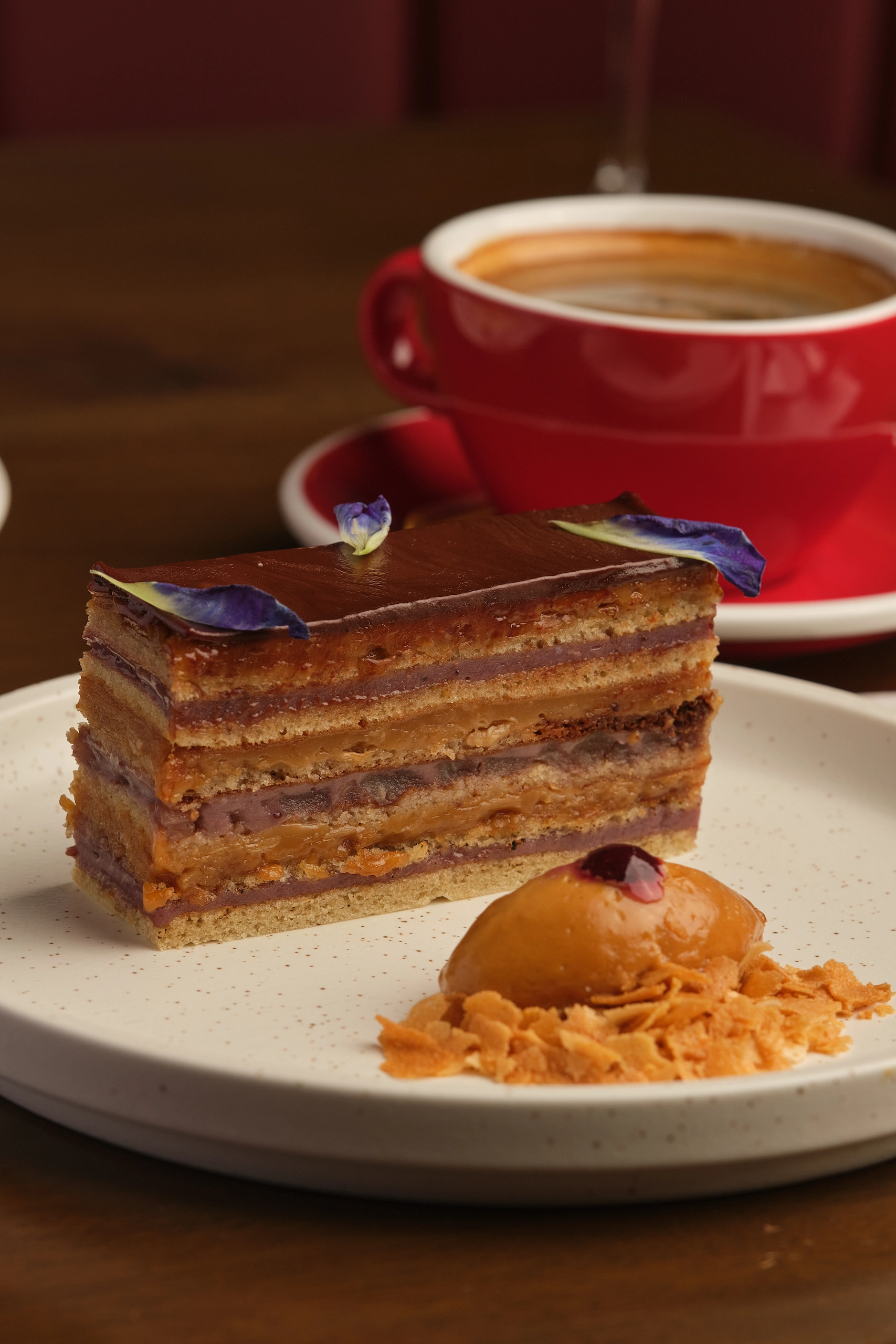 Slice of layered chocolate cake with glossy chocolate glaze and a small flower garnish on a white speckled plate.
