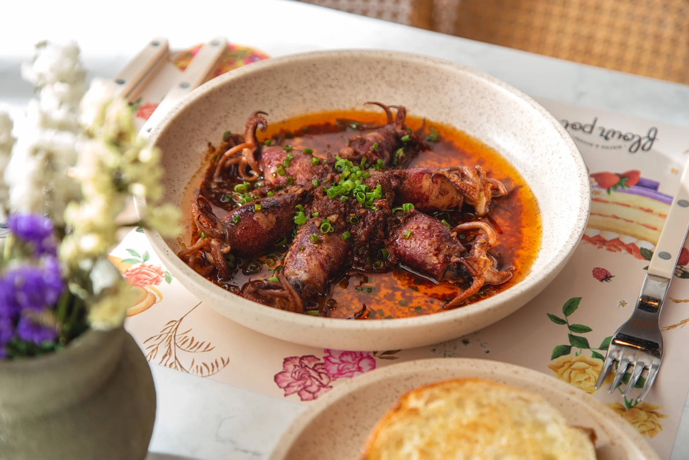 Flour Pot's Calamaretti. Bowl of spicy squid in red chili oil, sprinkled with chopped green onions on a speckled cream bowl.