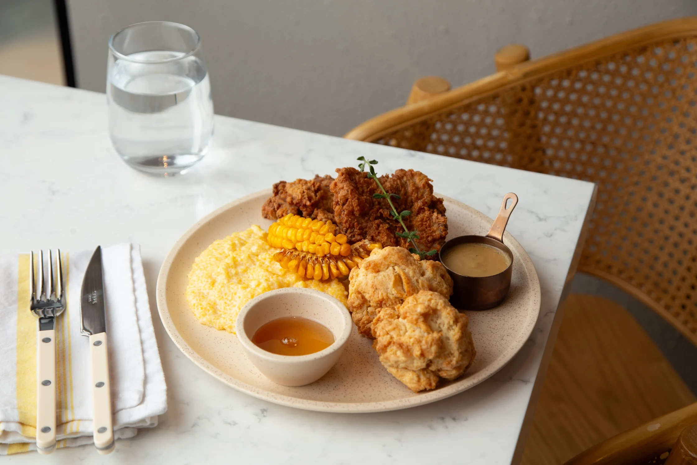 Flour Pot's Chicken and Biscuits. Plate with fried chicken, corn on the cob, fluffy mashed potatoes, biscuits, and a small cup of gravy with a side sauce on a beige plate at a marble table.