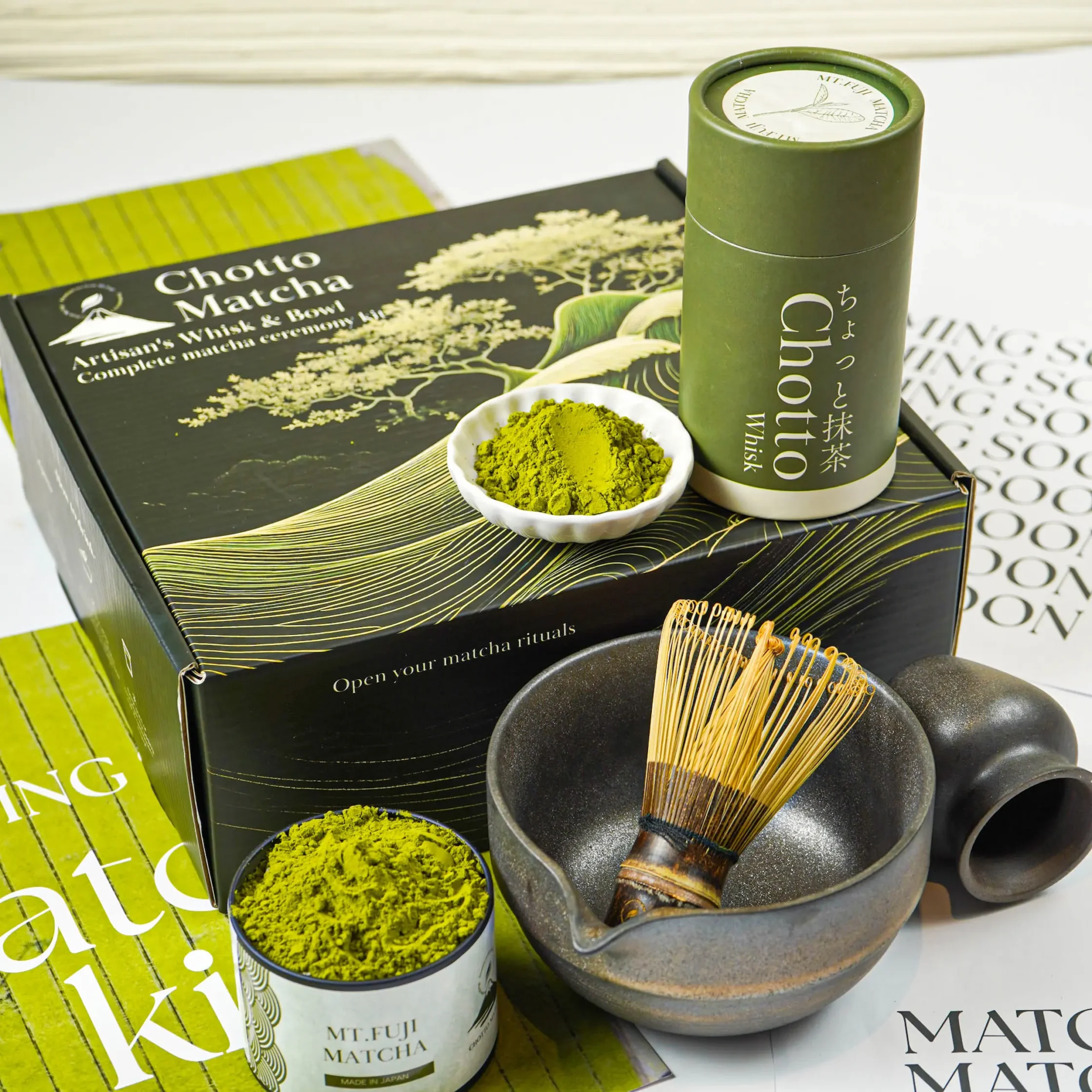 Close-up of a matcha tea kit: tins of green matcha powder, a bamboo chasen whisk, and black/green packaging labeled 'Chotto Matcha' for a Japanese tea ceremony kit.