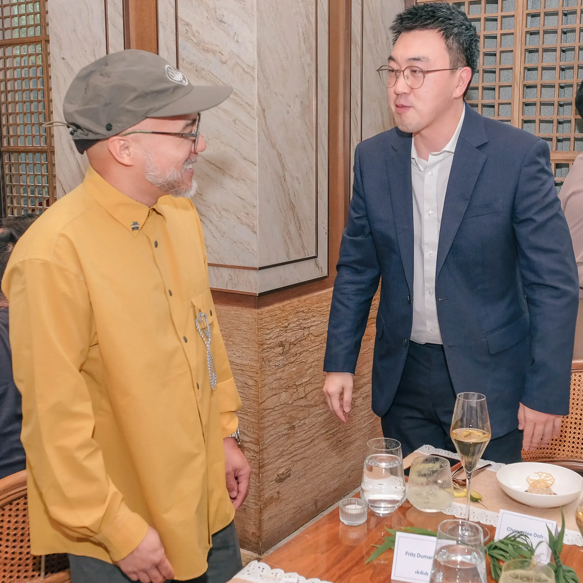 Two men in a restaurant talking near a table with drinks and plates on it.