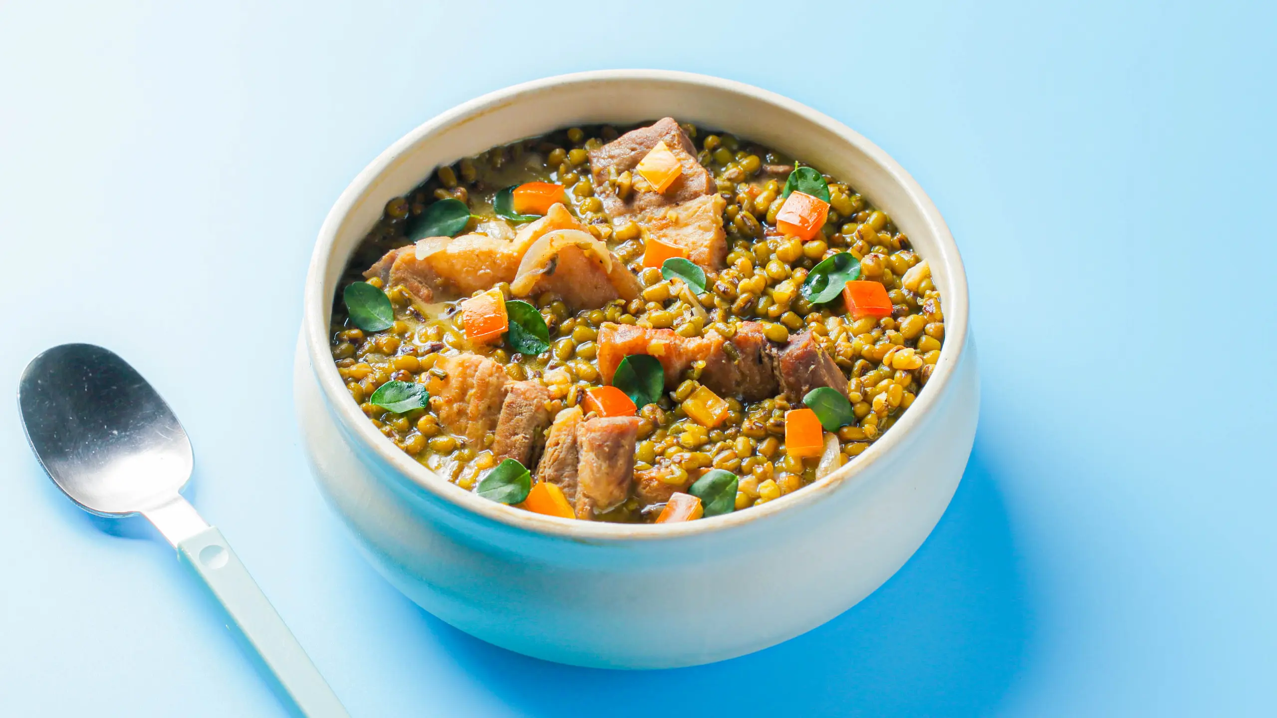 Bowl of sauteed munggo soup with tomatoes, pork chunks and greens on a blue surface, spoon nearby