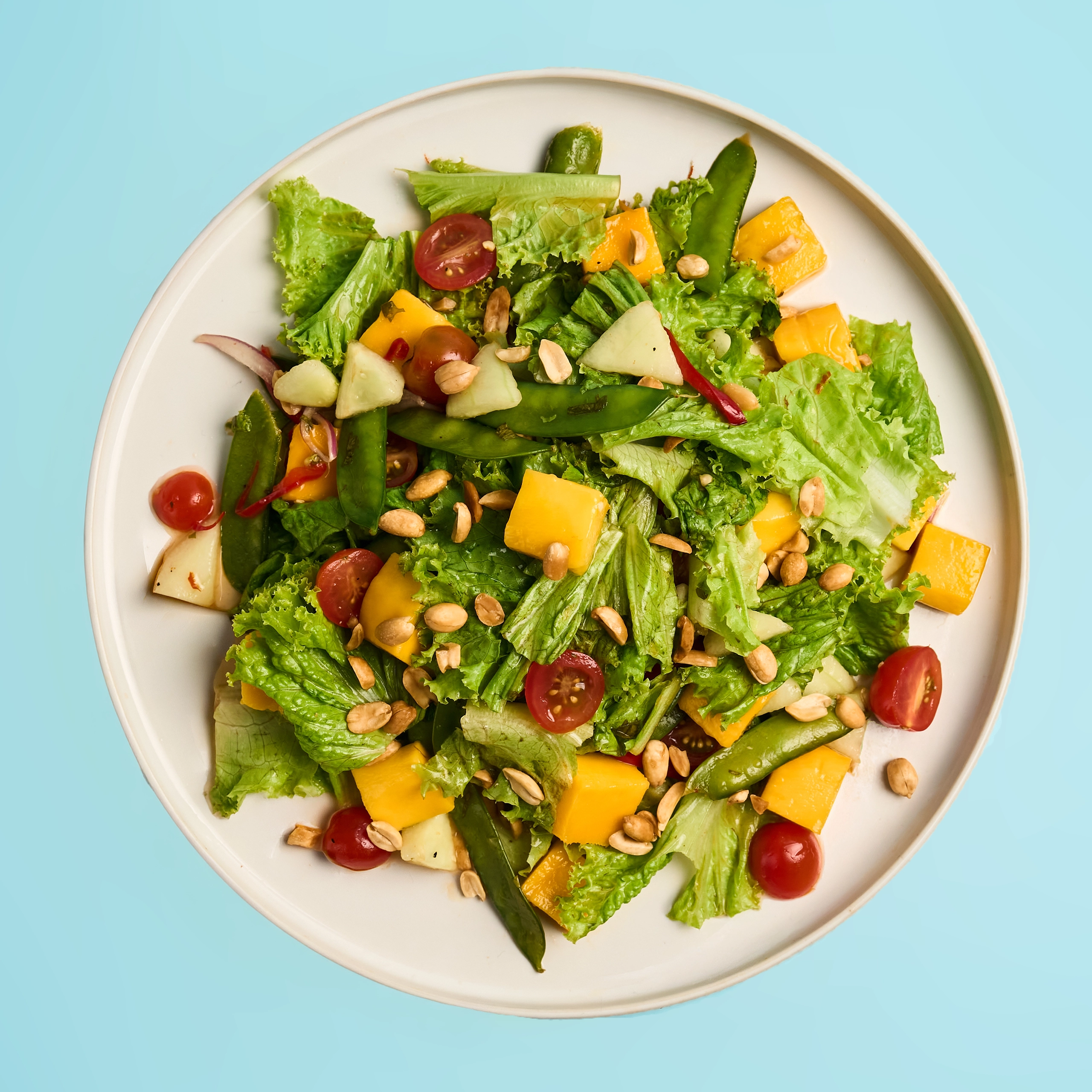 Plate of fresh mixed salad with lettuce, cherry tomatoes, mango chunks, cucumber, snap peas, and peanuts on a light blue background.