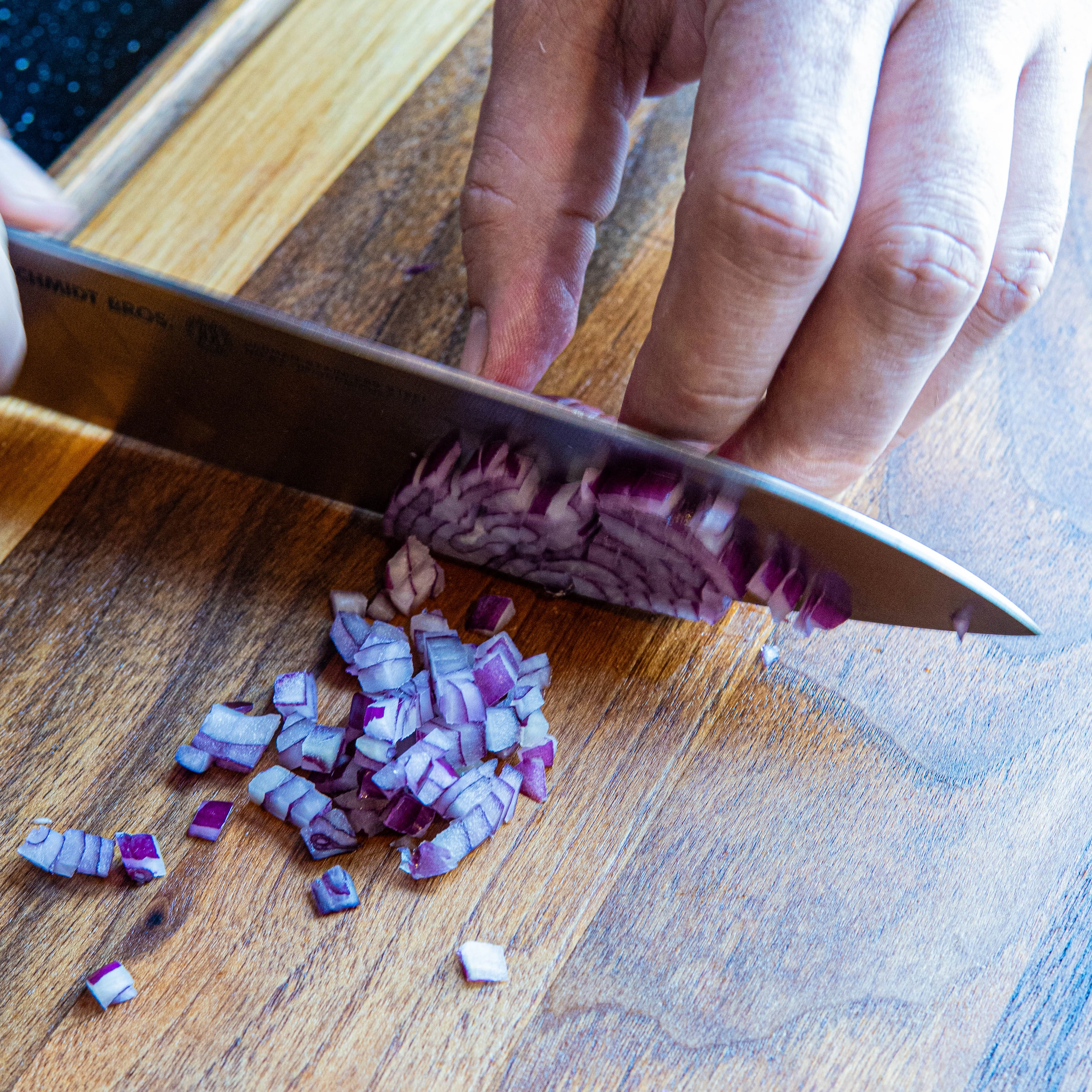 Person slicing a red onion on a wooden cutting board with a large knife, onion pieces gathered nearby.