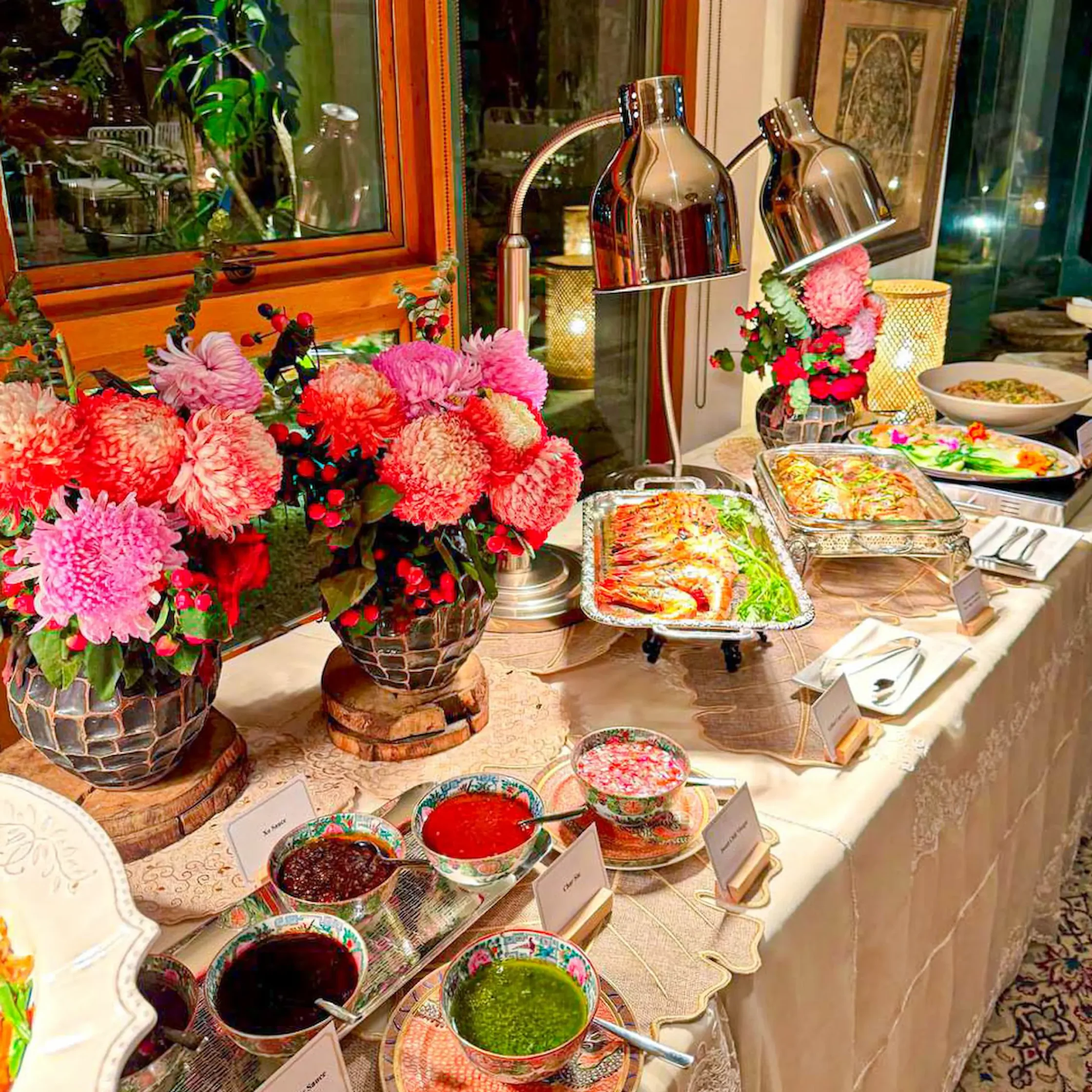 Colorful bowls of sauces on a decorated buffet table with pink flowers and ornate dishware near a window