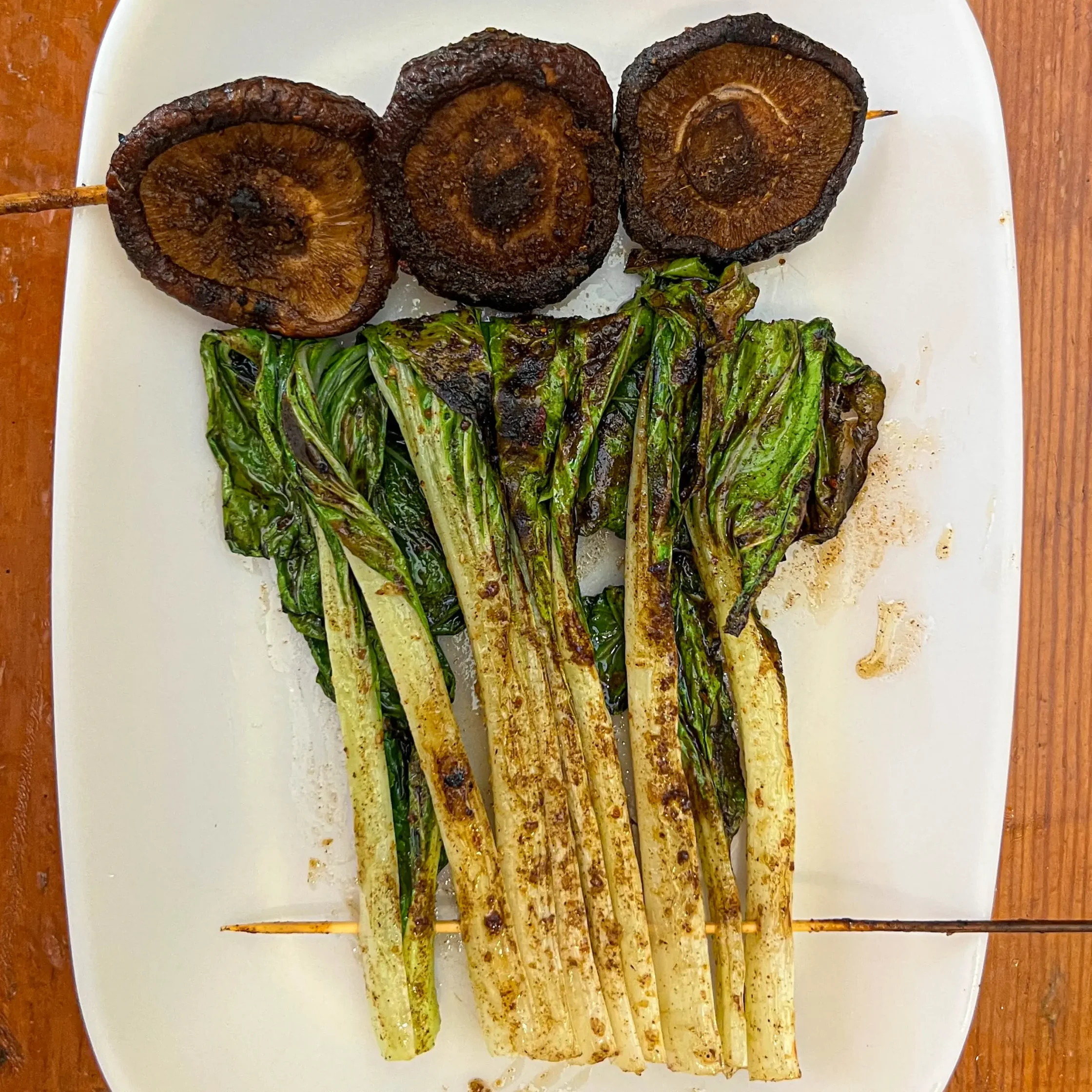 Skewered grilled mushrooms with charred mushroom caps over a bed of grilled asparagus on a white plate, viewed from above.