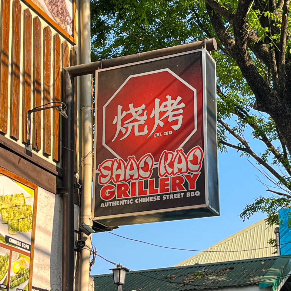 Red octagonal sign for a Chinese street BBQ restaurant mounted on a storefront next to a tree. (Est. 2013)