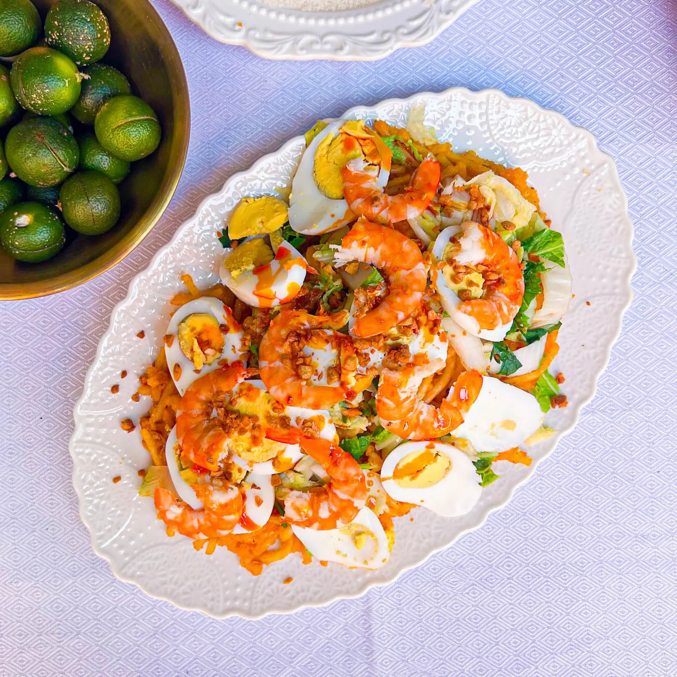 Pancit Malabon served on an oval white platter, featuring thick orange-tinted noodles topped with shrimp, sliced hard-boiled eggs, leafy greens, crispy garlic, and a savory orange sauce. Bowls of limes and a white granular ingredient are partially visible nearby.