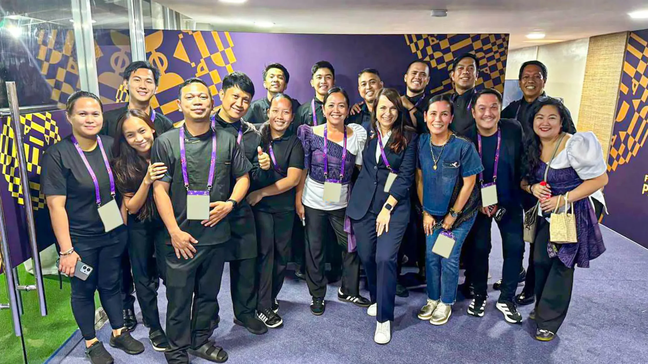 Group of people posing together at a corporate event hallway, wearing matching purple lanyards with badges.
