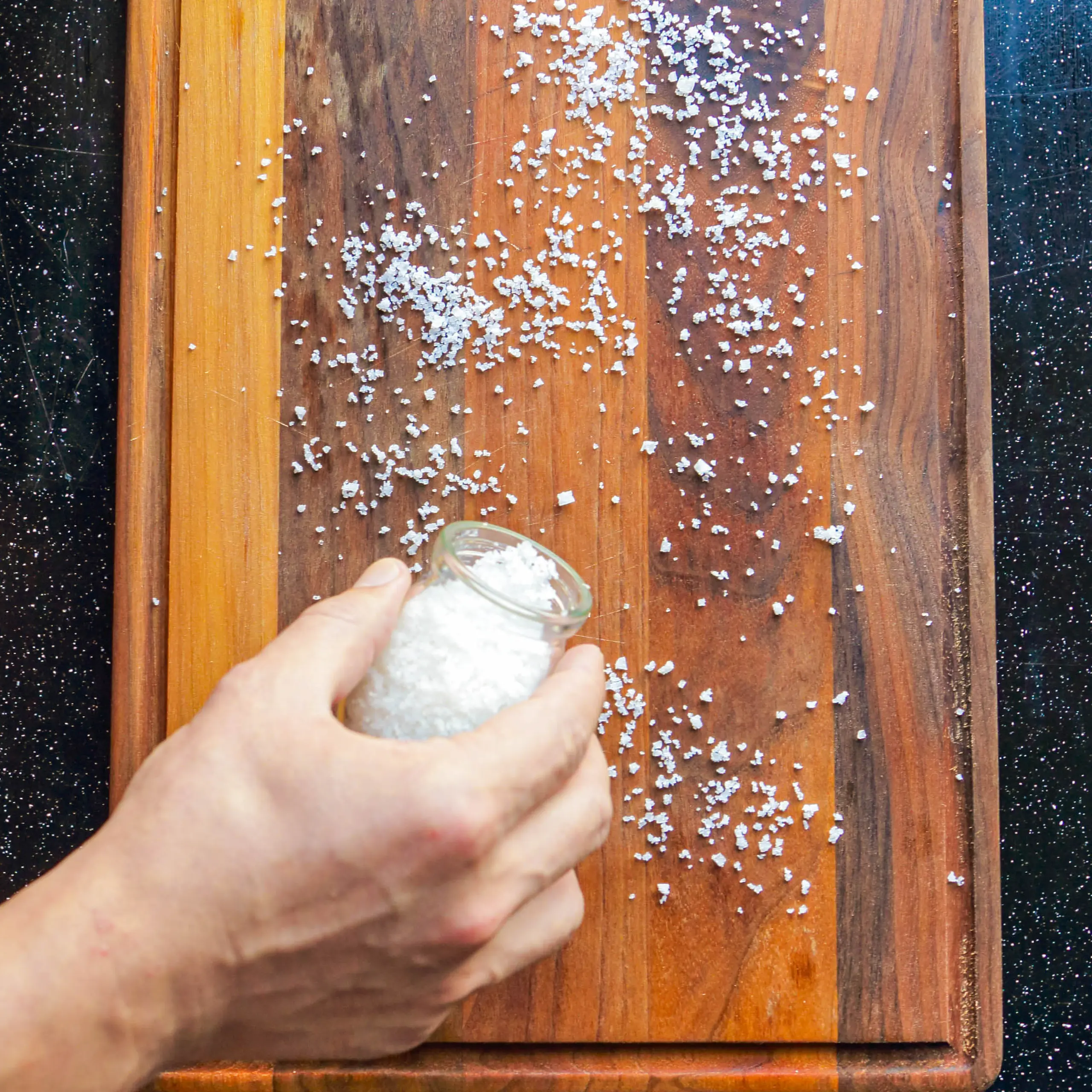 Hand sprinkling coarse salt from a glass jar onto a wooden cutting board, with salt crystals scattered around.