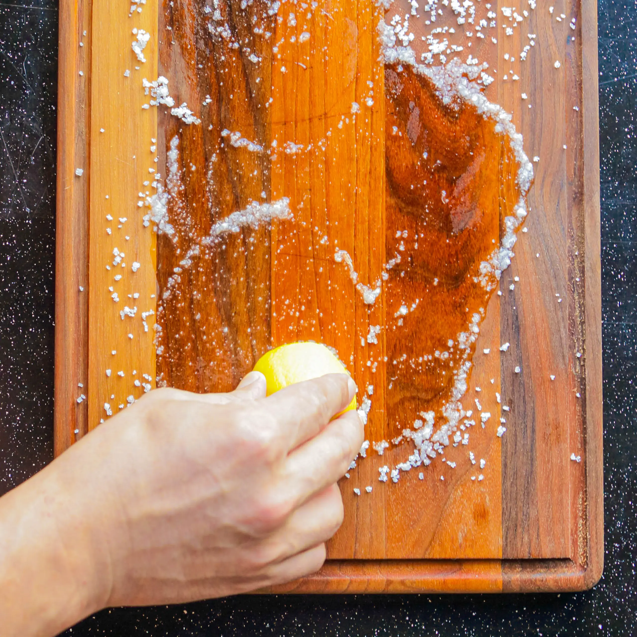 Hands squeeze a lemon over a wooden cutting board as coarse salt flakes scatter around the surface viewed from above, bright and tactile.