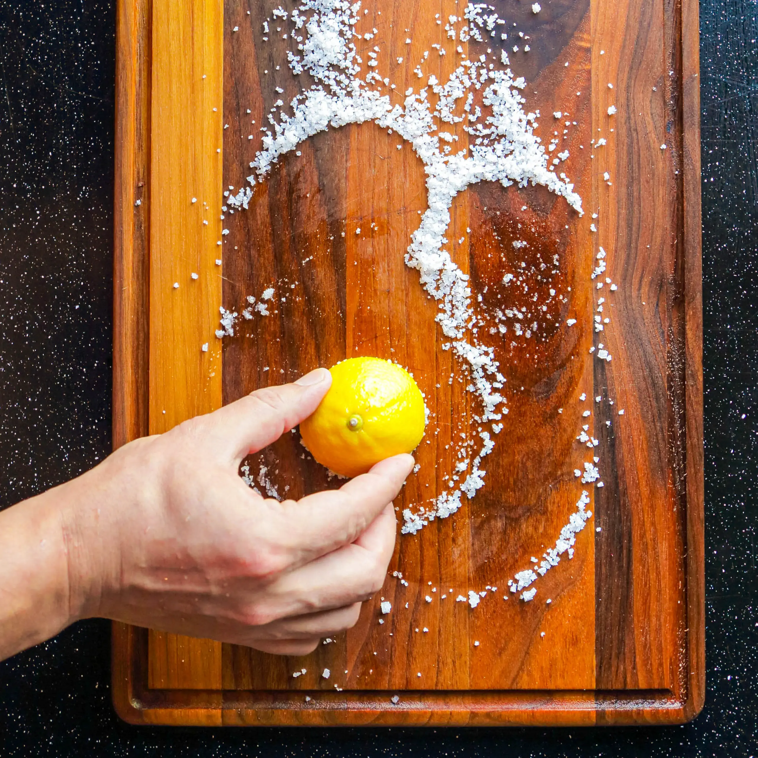 Hand holding a bright lemon over a wooden cutting board with a ring of coarse salt scattered around it.