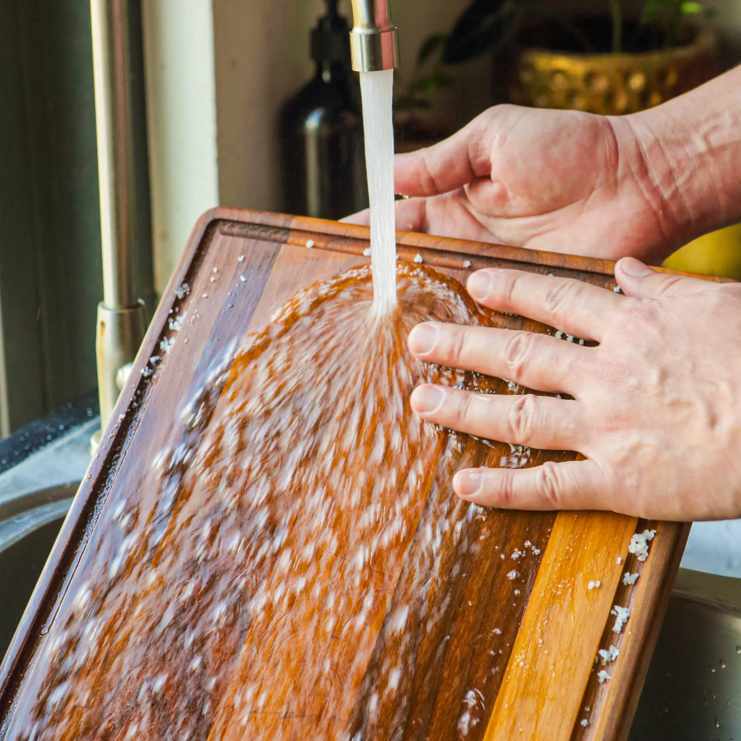 Person washing a wooden cutting board under a running faucet at a sink, water splashing on the board.