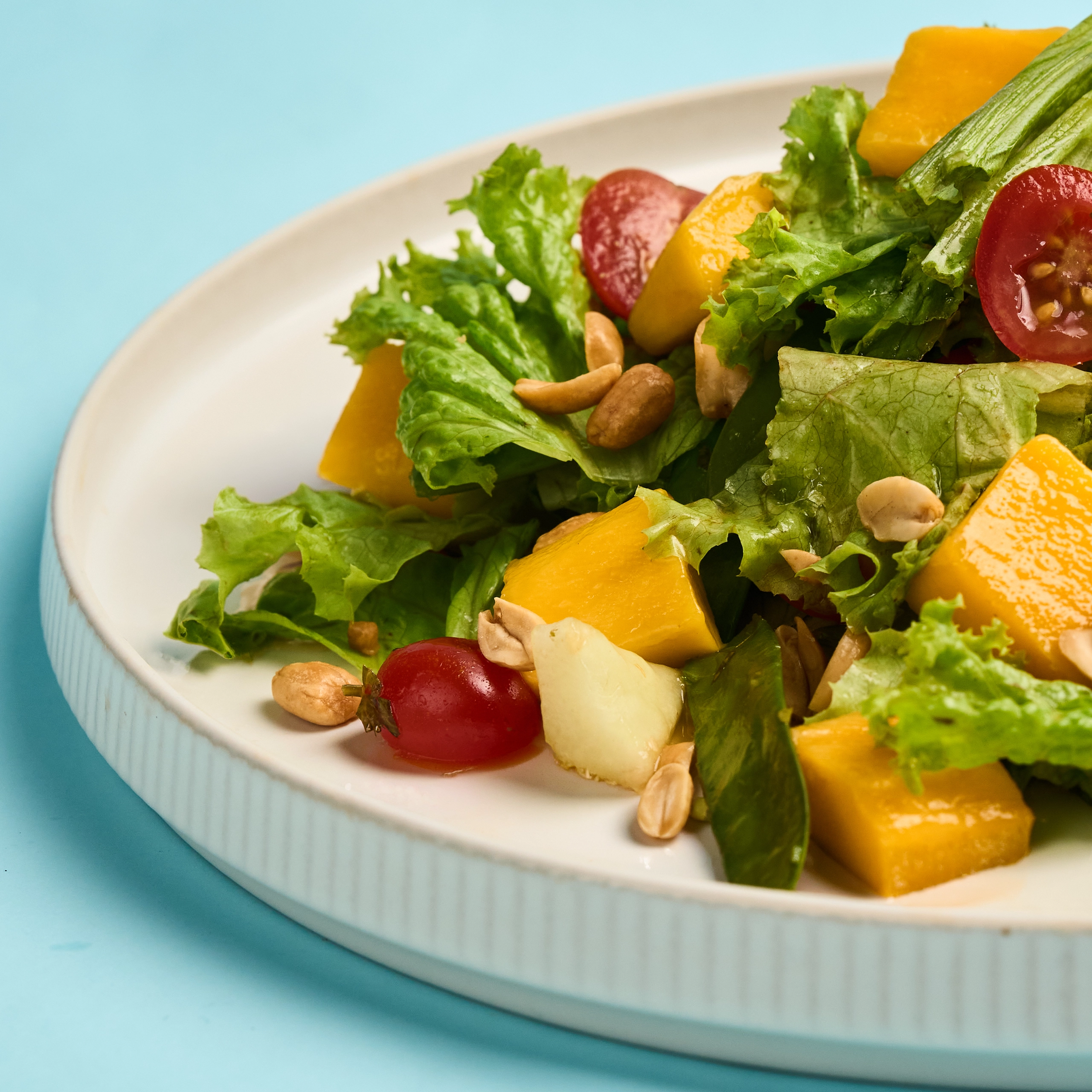 Close-up of a fresh salad in a white bowl with lettuce, mango chunks, cherry tomato halves, and peanuts against a light blue background.