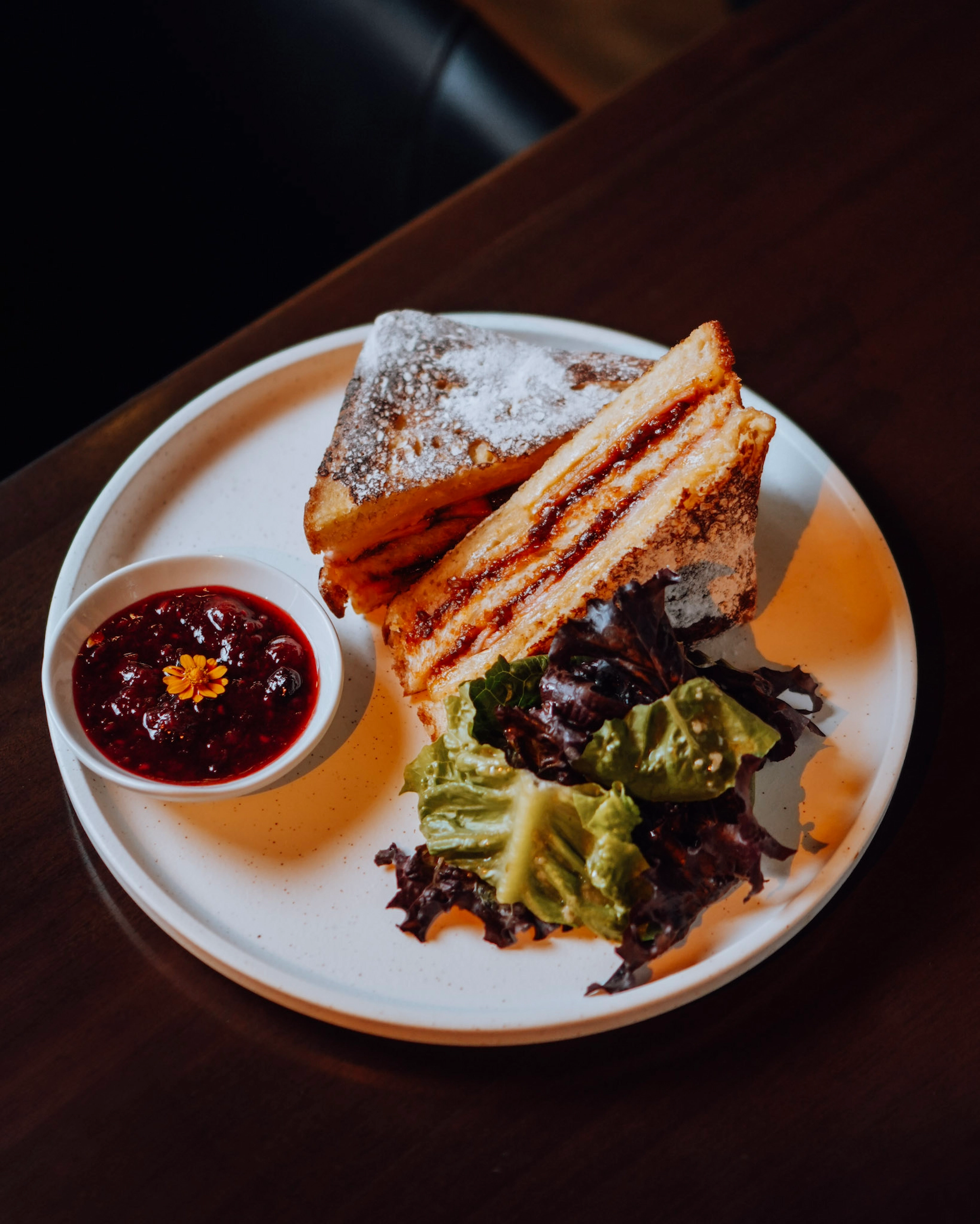 Triple-layer sandwich with jam, dusted sugar, and greens on a white plate, served with berry sauce on the side.