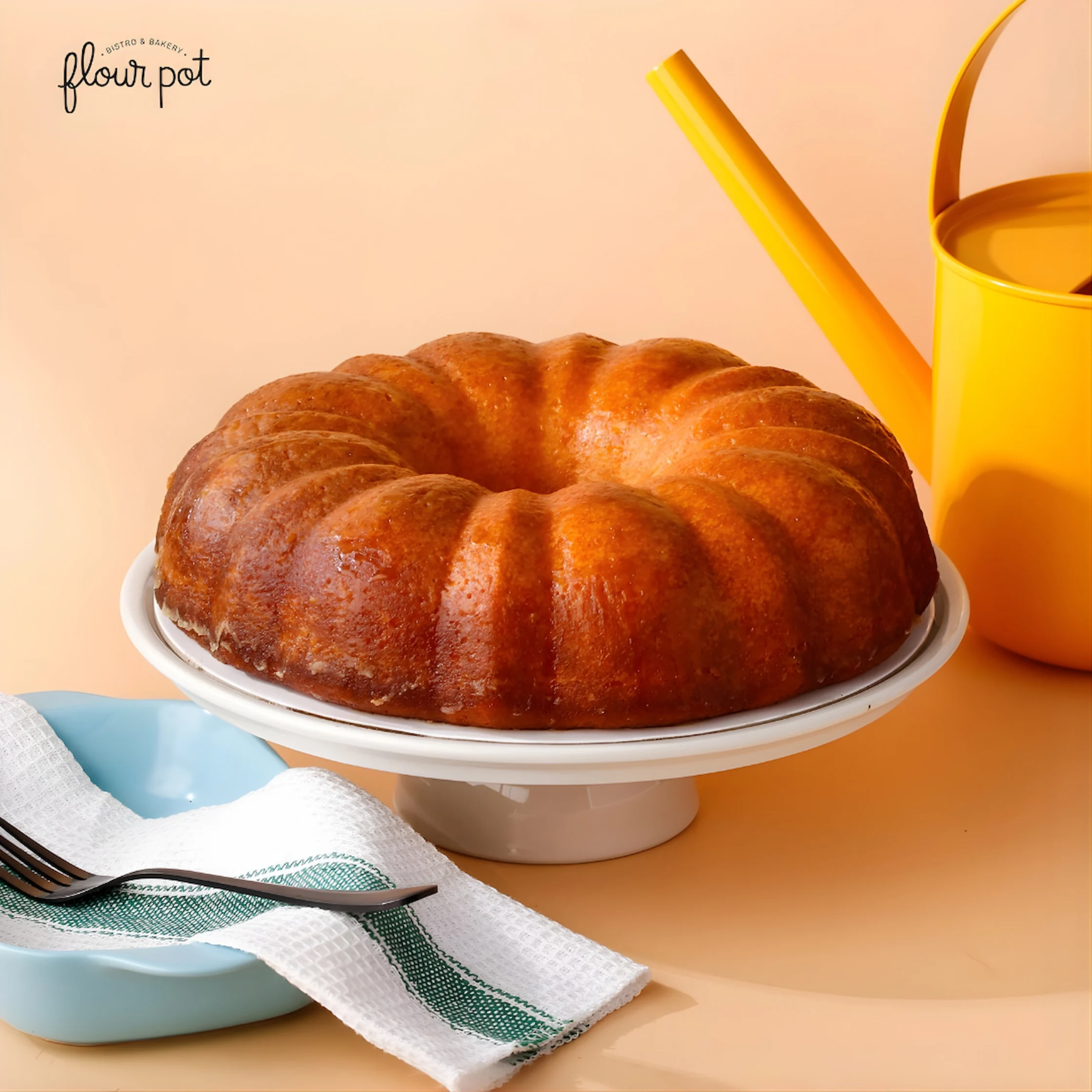 Golden-brown Bundt cake sits on a white pedestal plate with a blue dish, napkin, and fork in the foreground, yellow pitcher in the background.