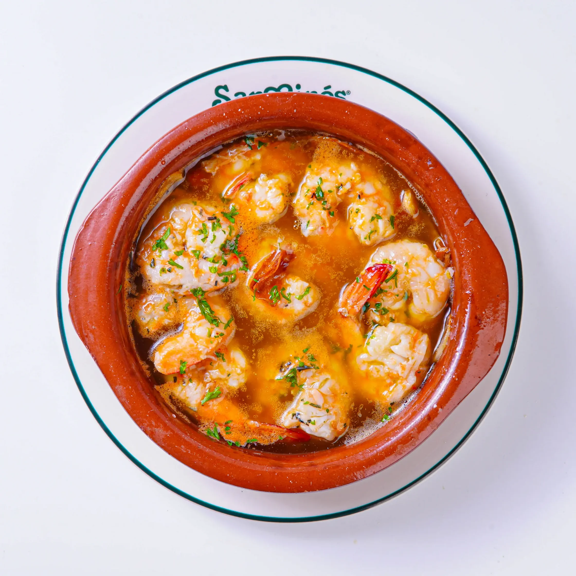 Shrimp in a tomato-based seafood stew, garnished with parsley, in a red ceramic pot on a white plate.