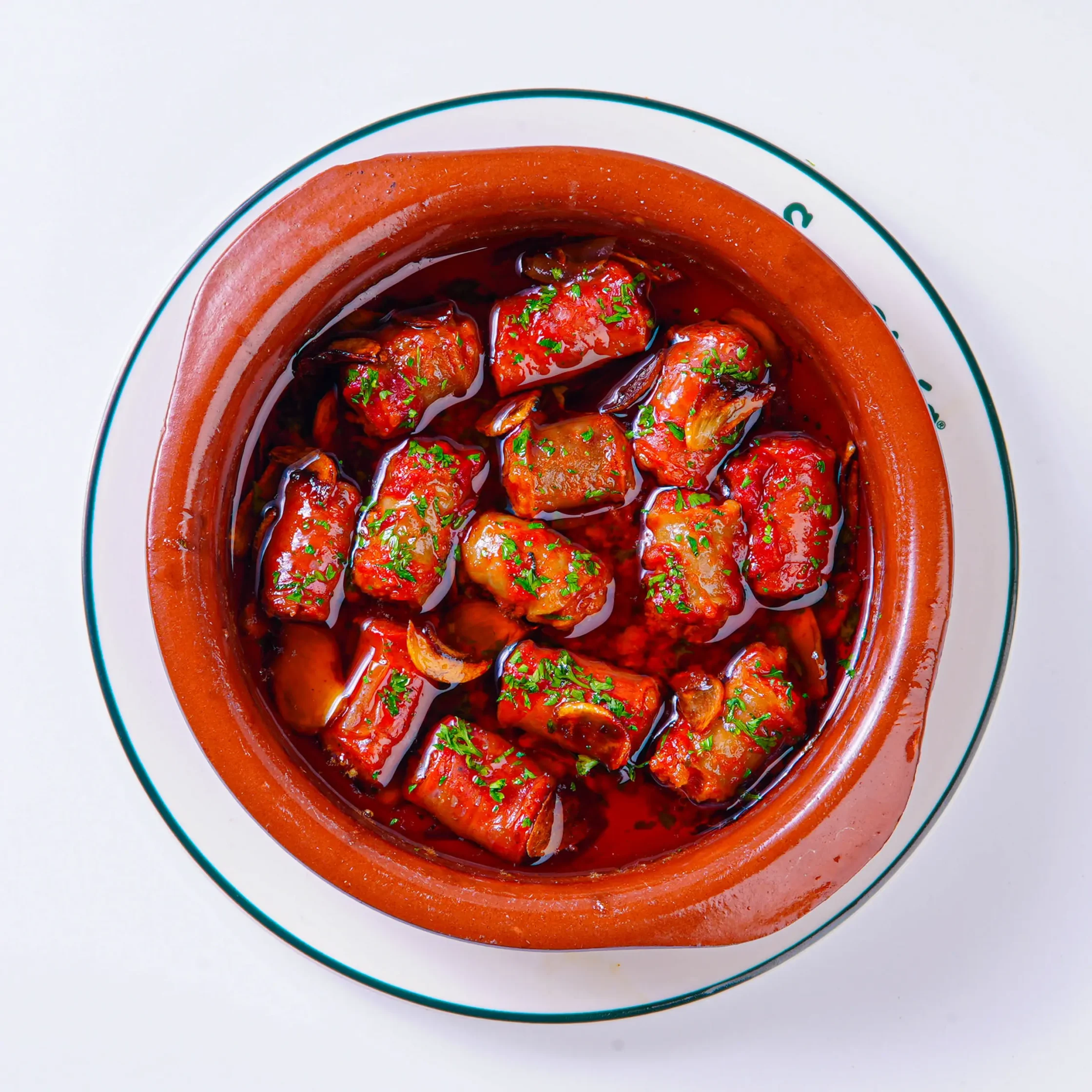 Braised meat chunks in bright red tomato sauce in a red ceramic pot, topped with chopped parsley.
