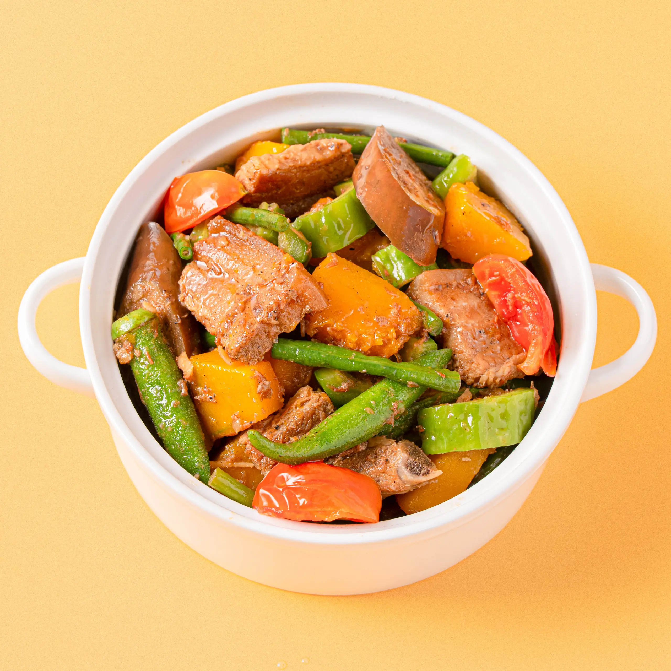 Stew with beef, bell peppers, green beans, and tomatoes in a white pot on a yellow background