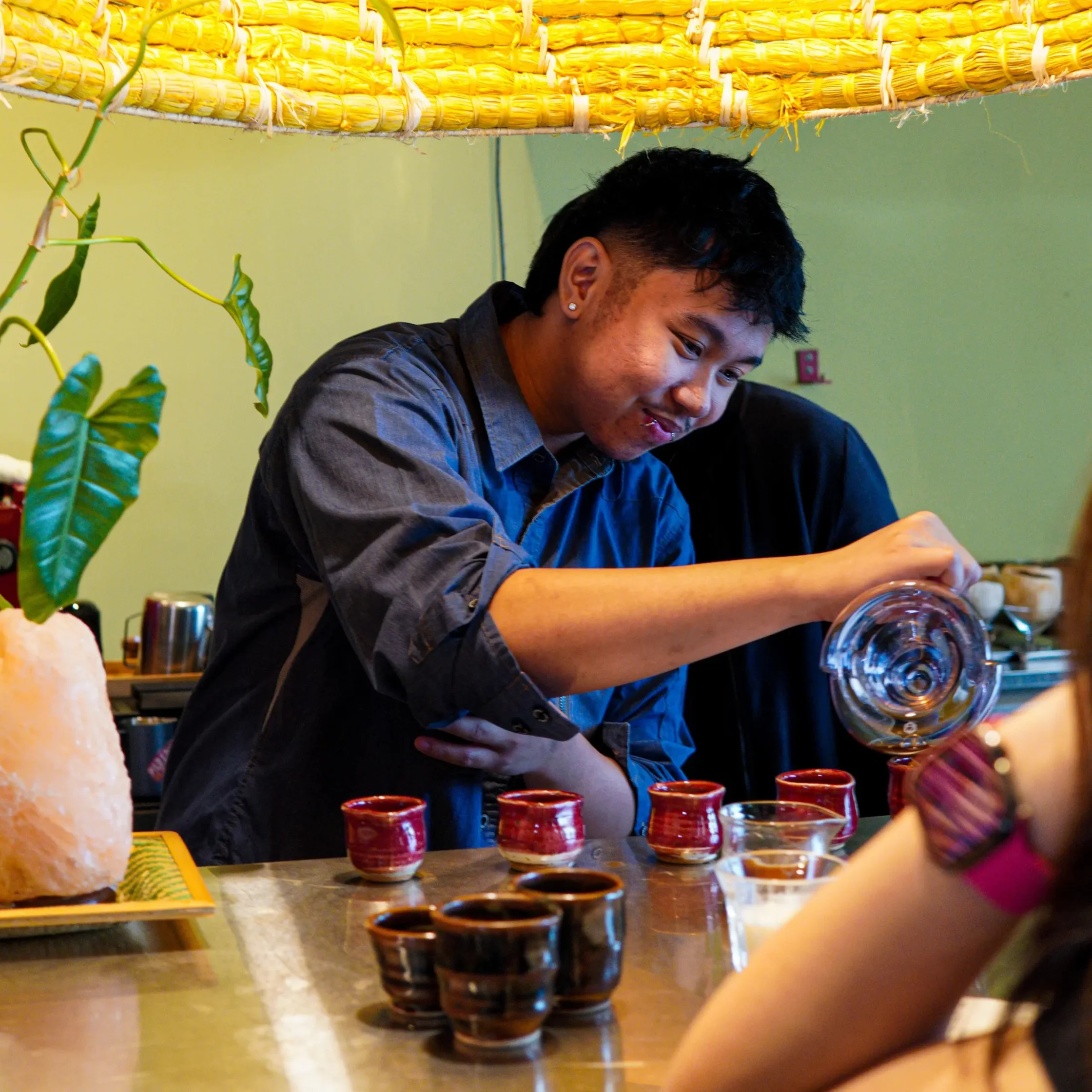 Man in a blue shirt pours a drink from a glass pitcher into small ceramic cups on a counter at a bar or cafe.