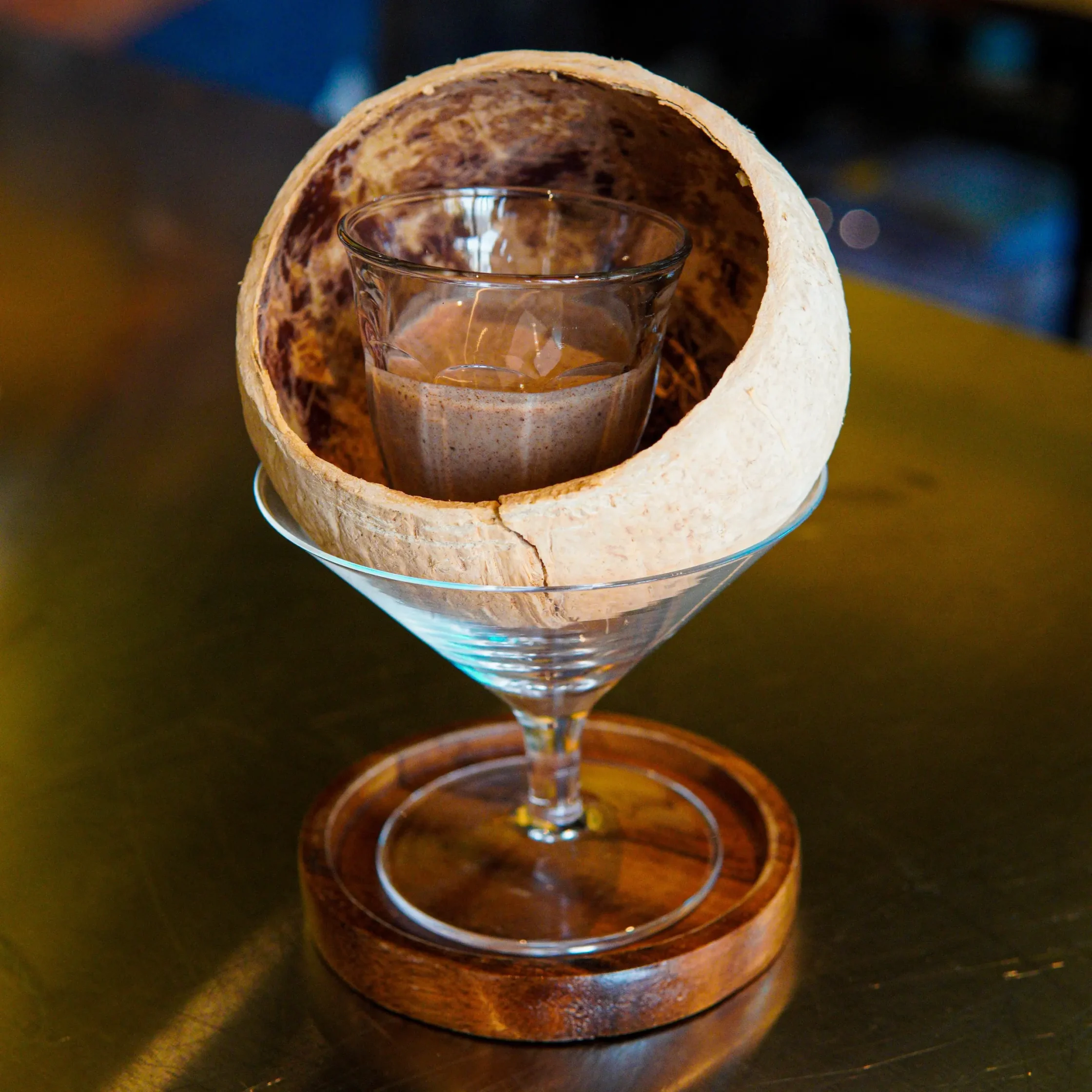 Cocktail served in a martini glass inside a cracked coconut shell on a wooden coaster.