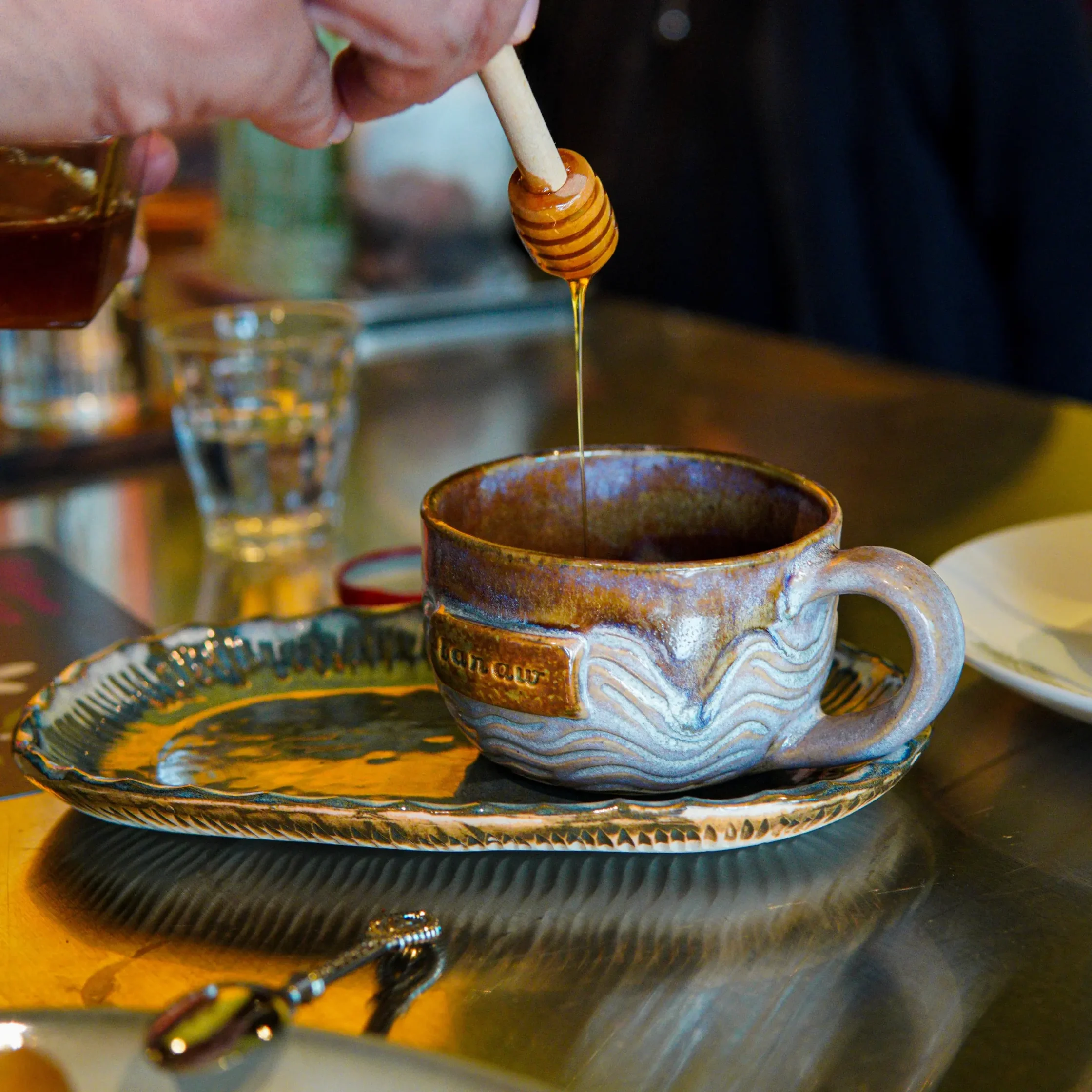 Hand pouring honey from a wooden dipper into a brown ceramic mug on a decorative plate.