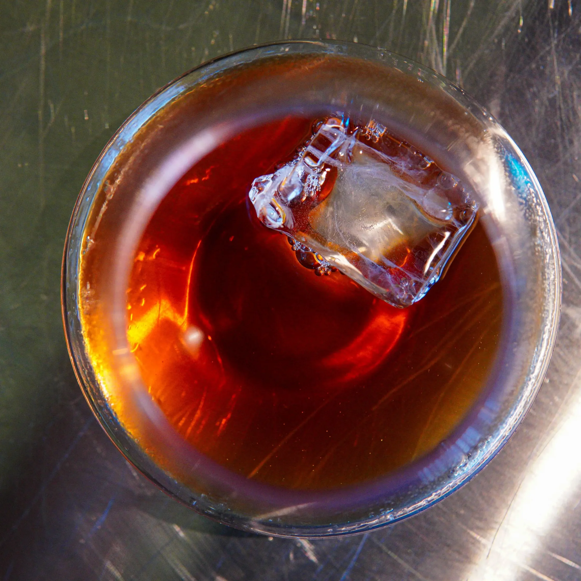 Top-down view of a glass of iced coffee with a large ice cube melting in it, on a metallic surface.