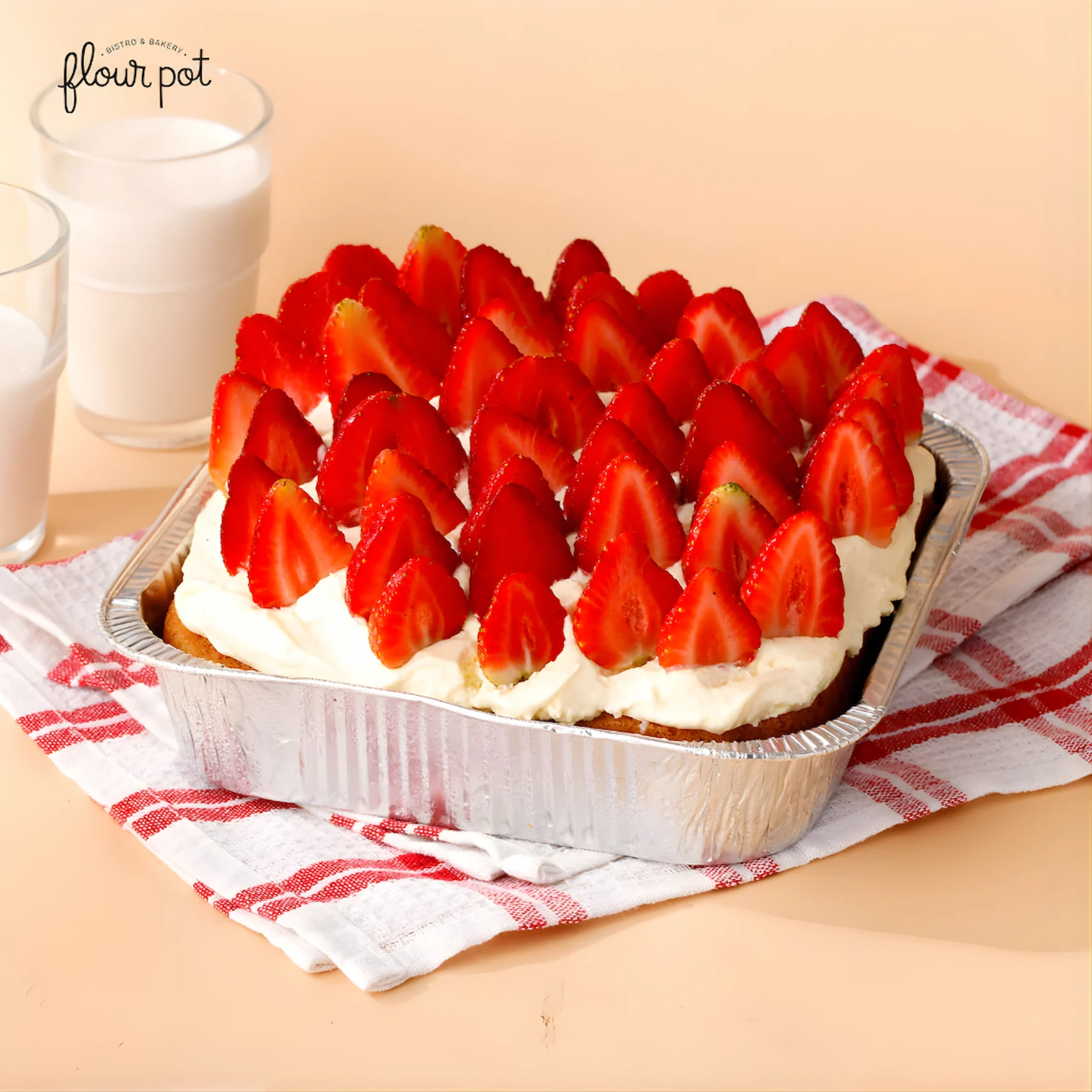 Flour Pot's Tres Leches. Strawberry-topped cake in a foil pan with whipped cream, set on a red-checked cloth beside glasses of milk.