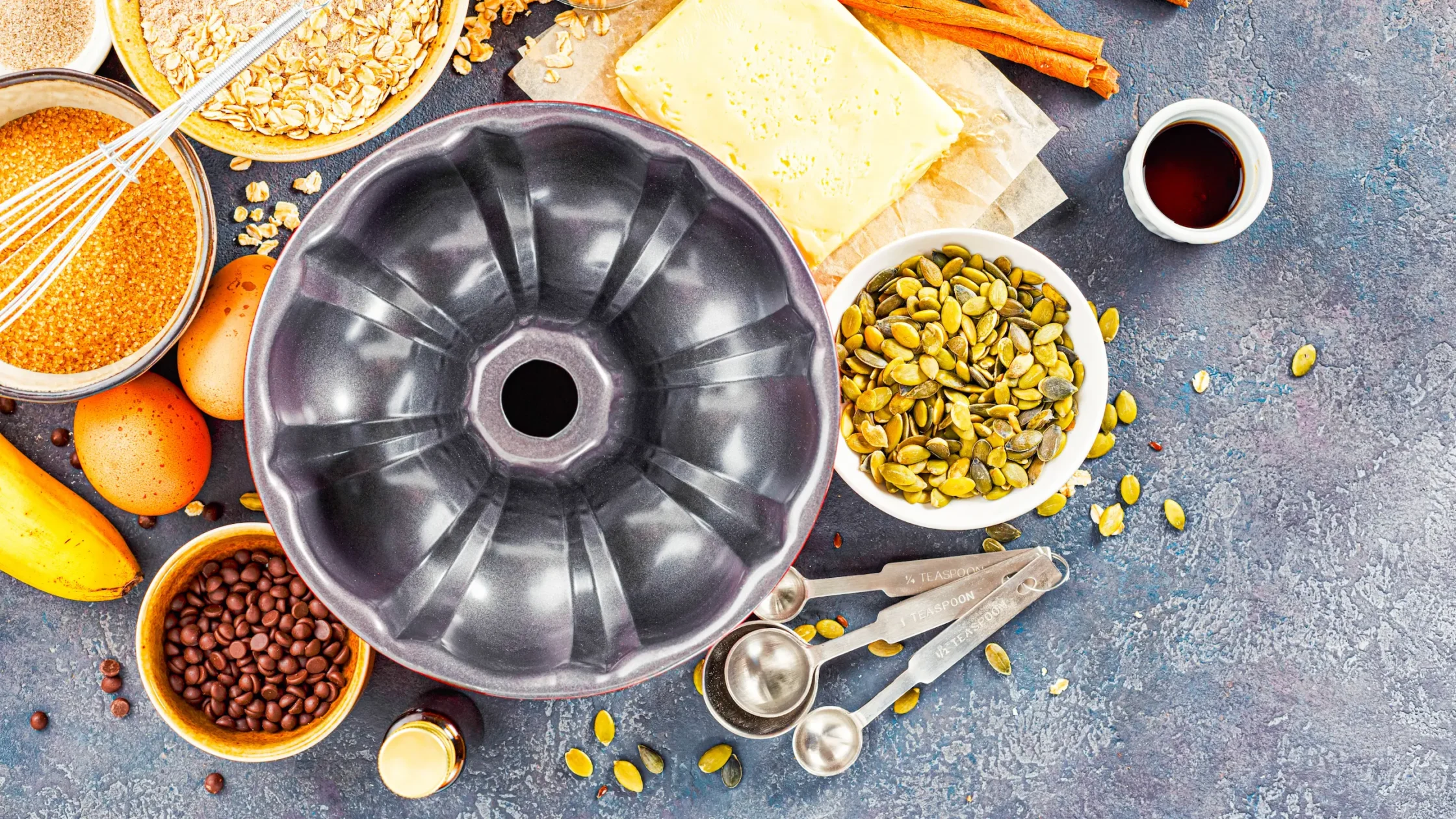 Top-down view of baking ingredients arranged around a Bundt pan: butter, eggs, brown sugar, oats, pumpkin seeds, spices, and measuring spoons.