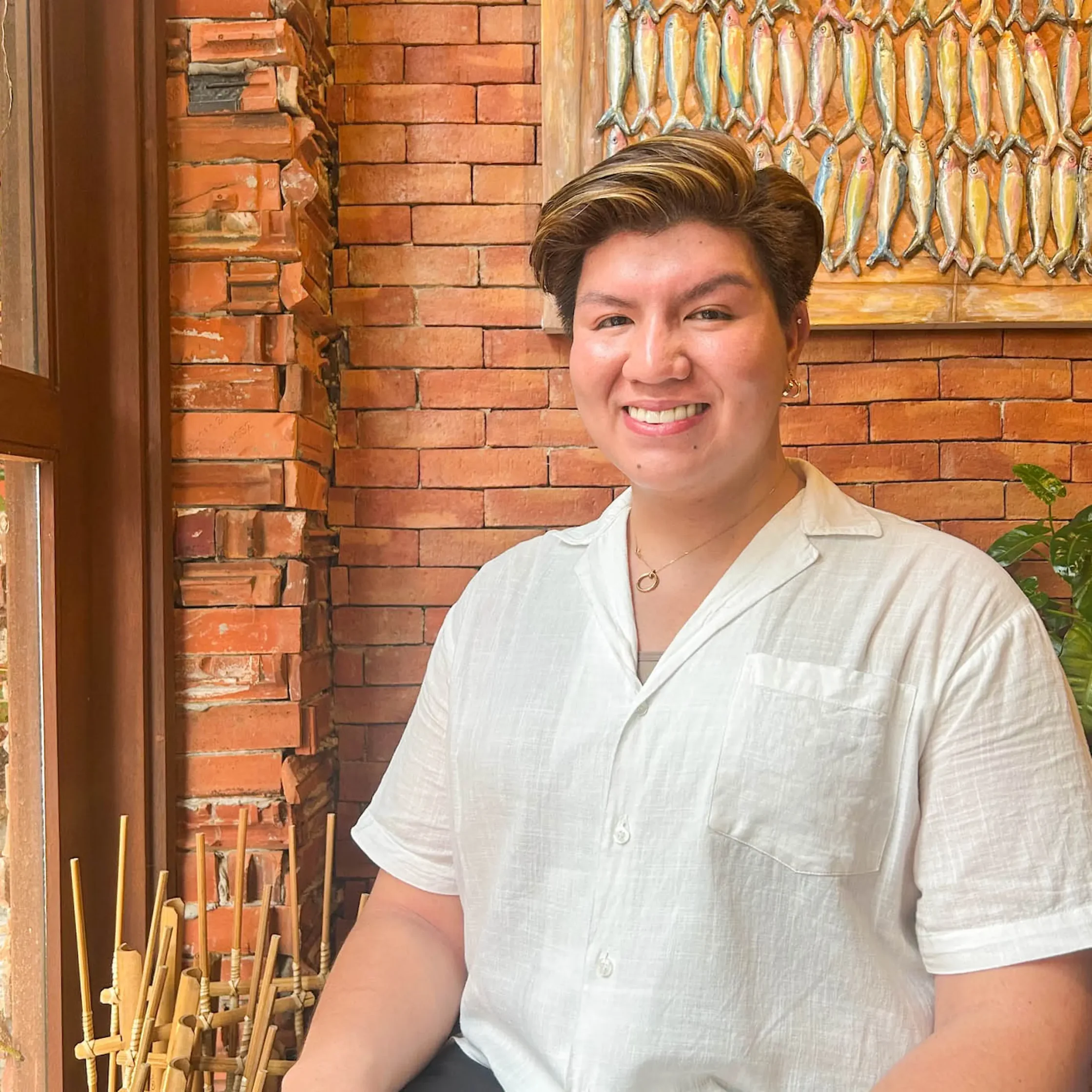 Smiling person facing the camera, wearing a light shirt, standing in front of an orange brick wall with wooden sticks in the foreground and a row of colorful wall decorations behind.