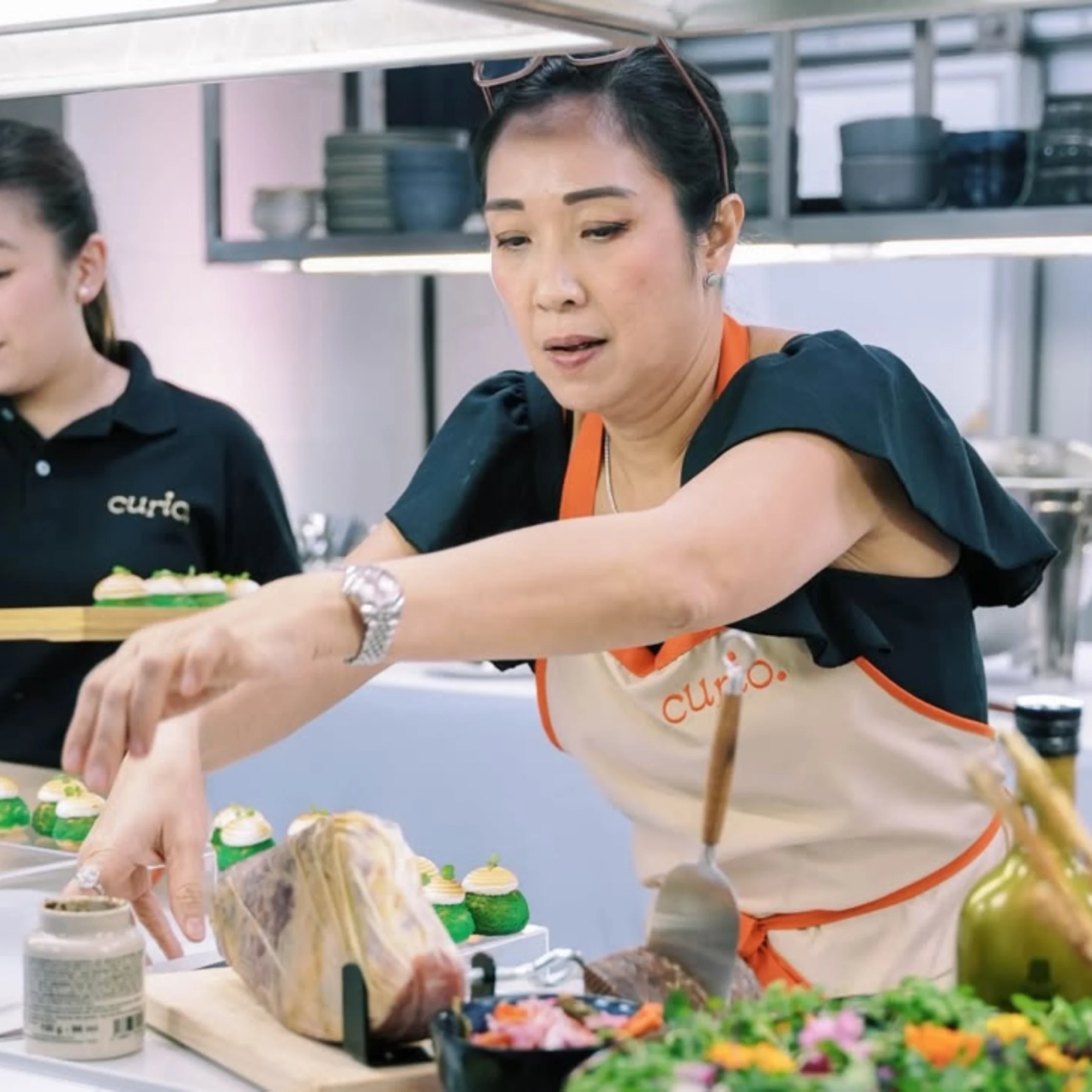 Bakery worker in an orange apron arranges colorful cupcakes on a display tray in a bright kitchen.