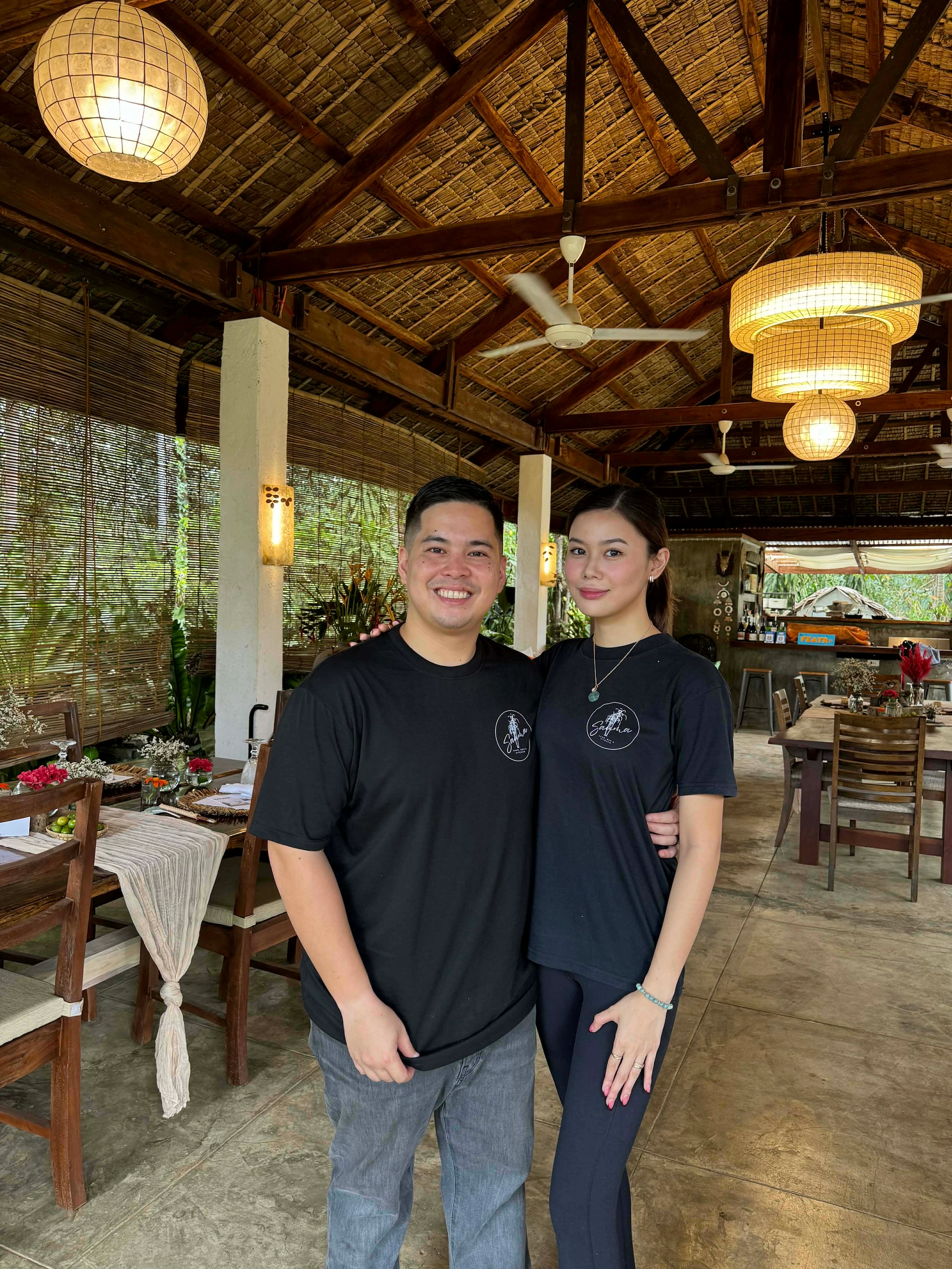 A smiling couple wearing black shirts standing arm in arm inside a rustic open-air dining area with wooden beams and wicker lamps.