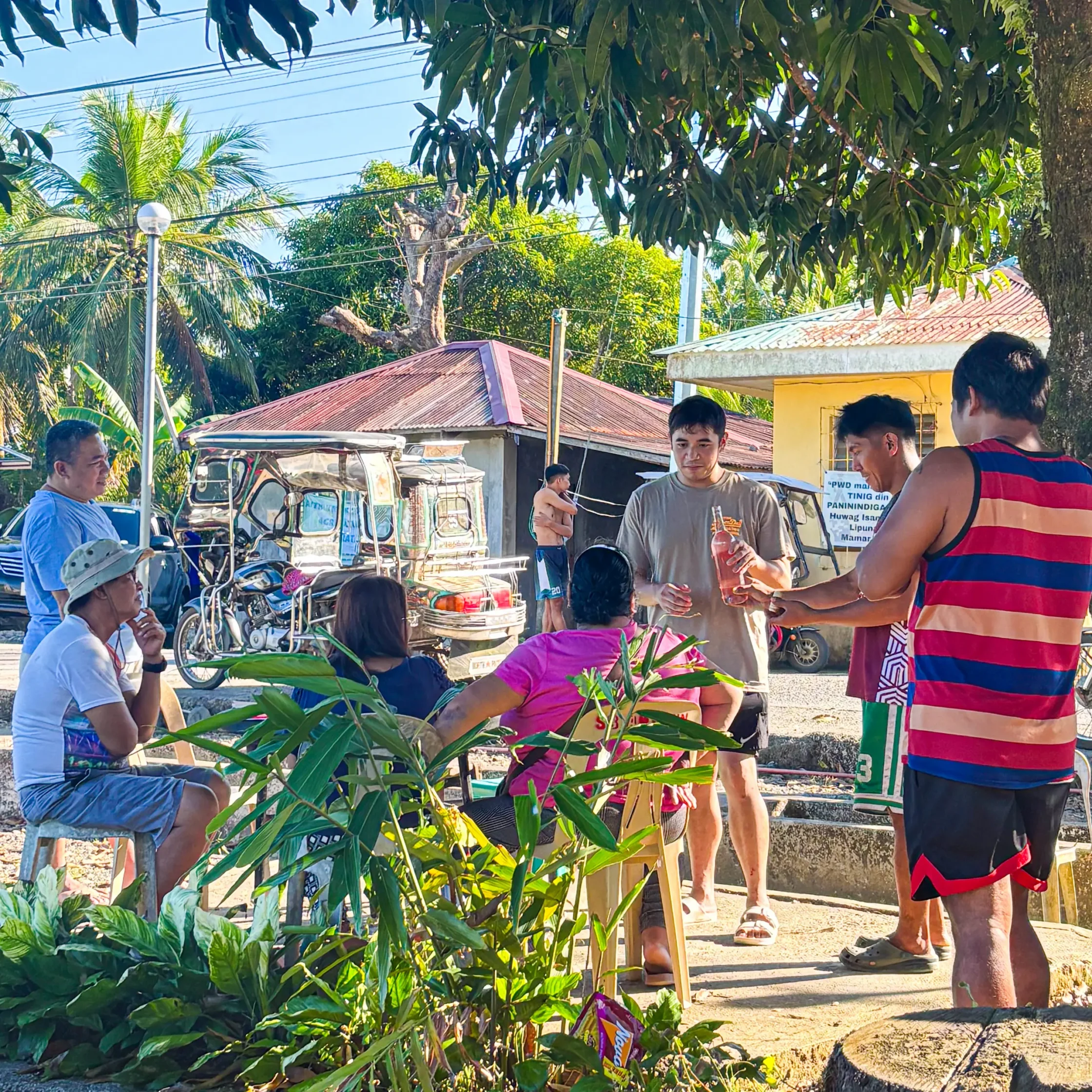 Group of people gathered outdoors among tropical plants and a tree, talking and holding drinks on a sunny day.