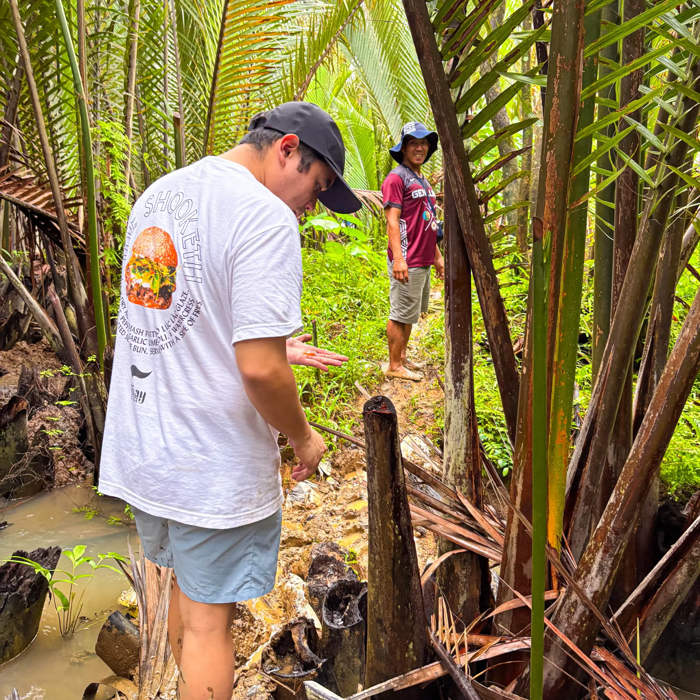 Two men in a tropical palm forest; foreground man in a white shirt and black cap looks down, background man in a maroon shirt and hat smiles.