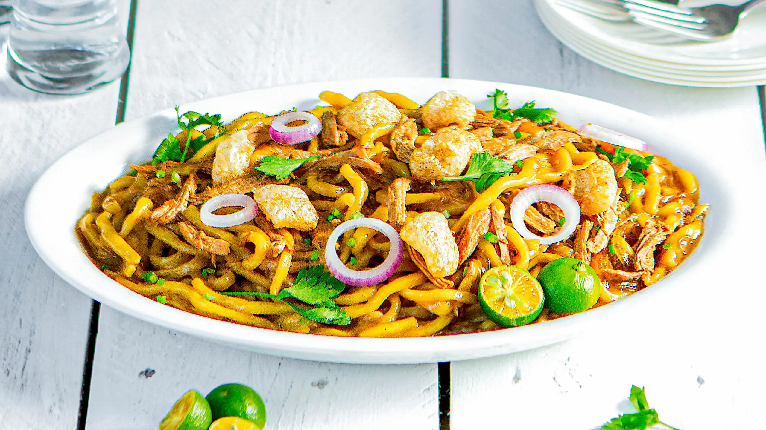 Plate of saucy pasta with yellow-orange sauce, garnished with red onion rings, crispy toppings, cilantro, and lime wedges.