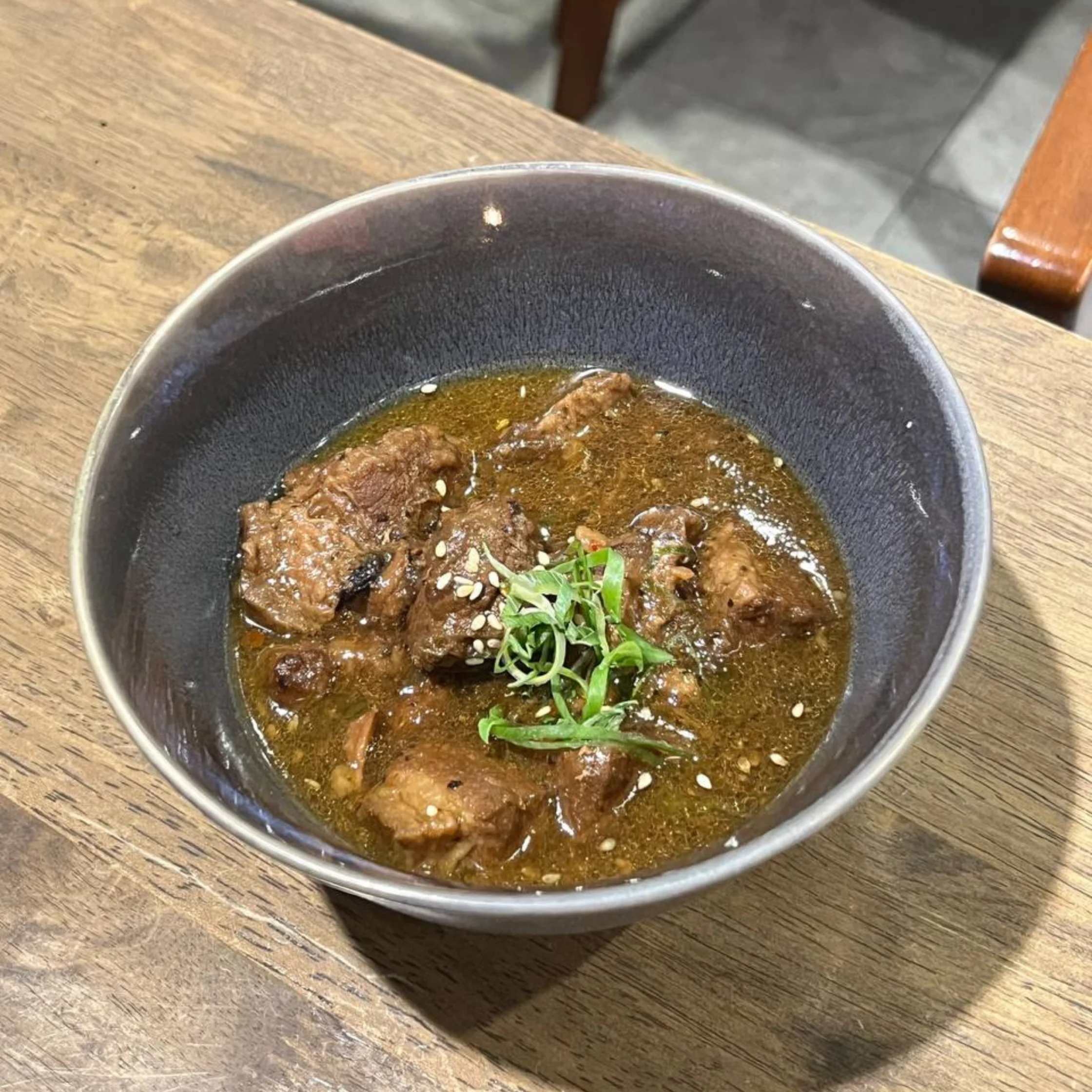 Beef stew in a dark bowl topped with greens and sesame seeds on a wooden table.